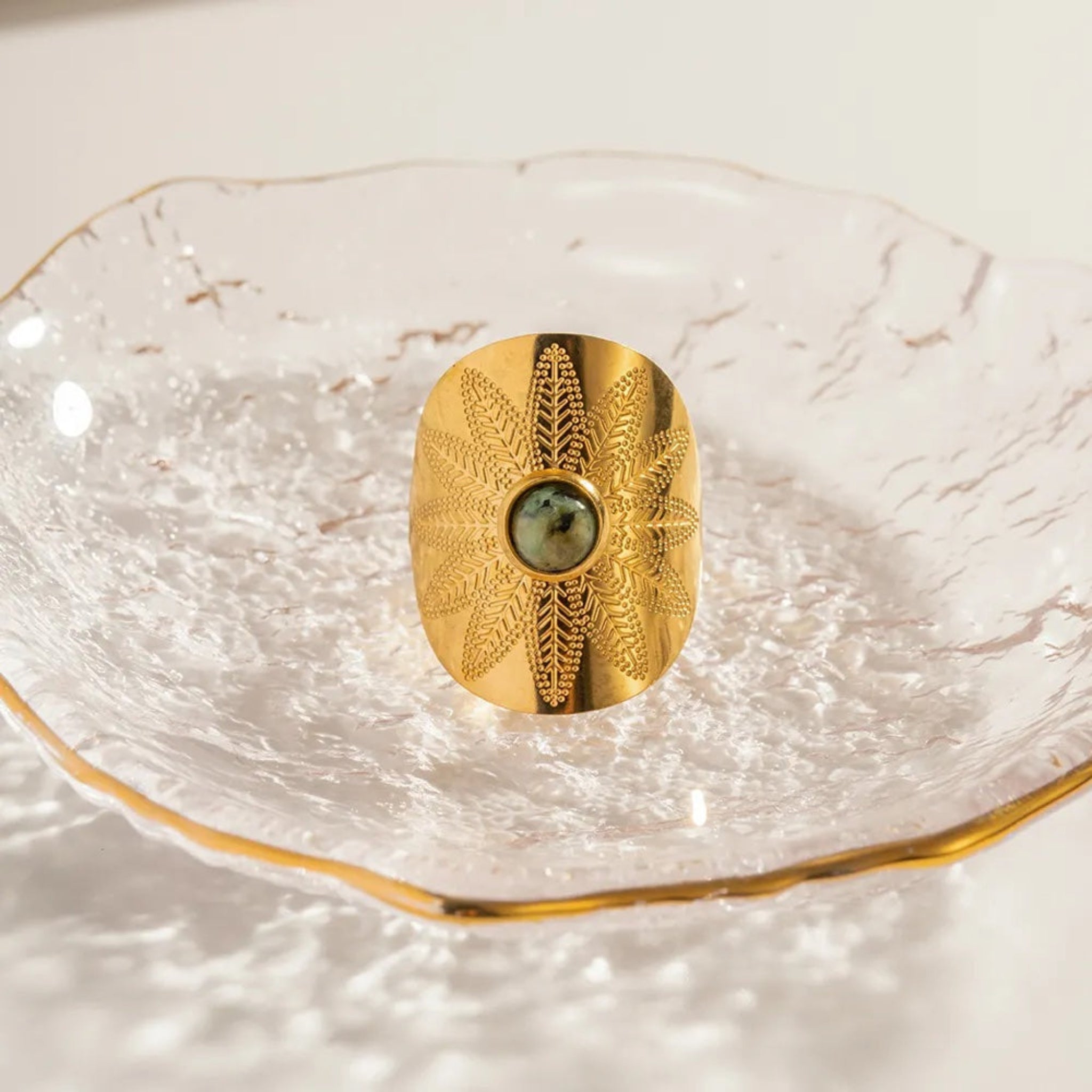 A gold-colored ring with a central African Turquoise gemstone, placed on a clear glass dish.