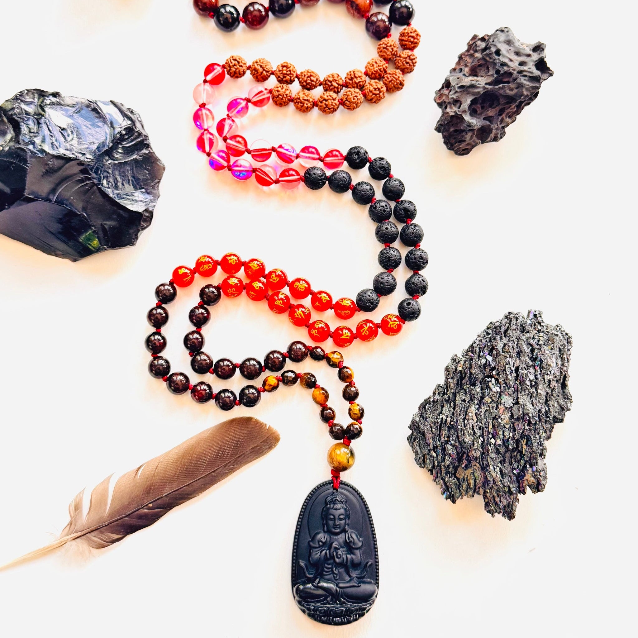 A variety of beaded necklaces laid out with different beads including red, black, and multicolored beads, along with a black obsidian Buddha pendant, surrounded by natural stones and a feather.