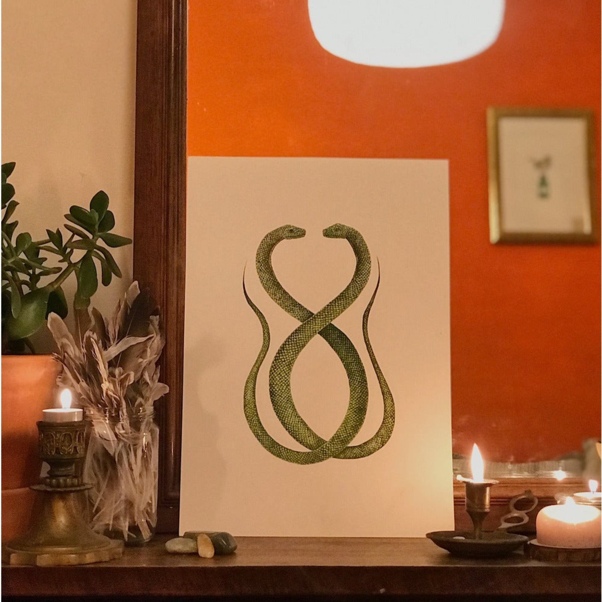Decorative wall art with two green snakes on a beige background, placed on a wooden surface with candles and plants.