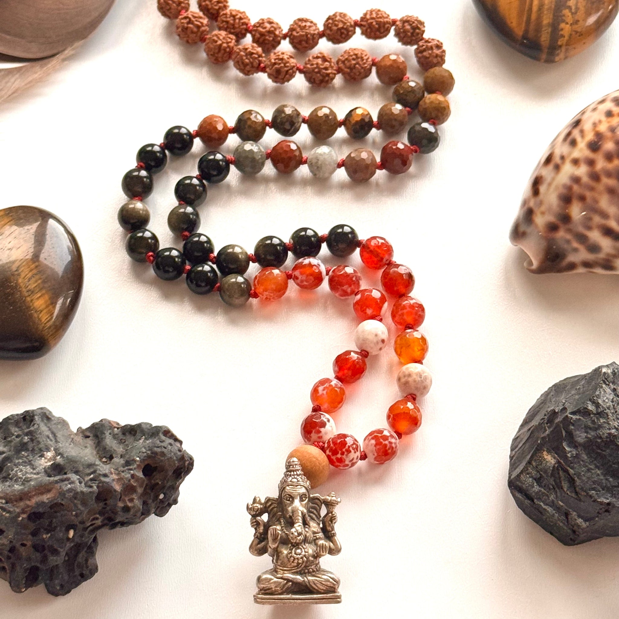 Necklace with various beads and a statue, surrounded by stones and shells on a white background