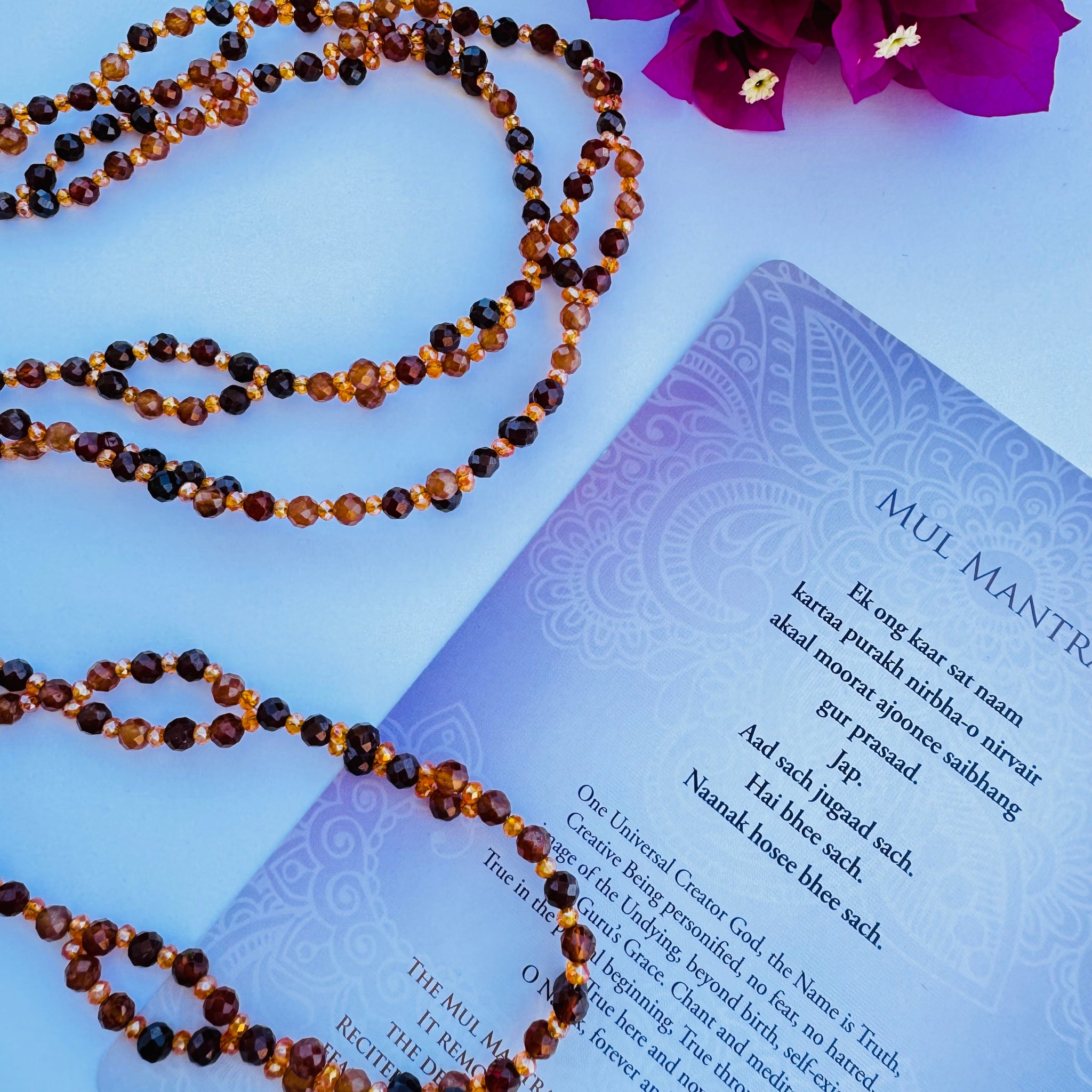 Beaded brown and orange tantric necklace on white surface decorated with pink flower and mul mantra card
