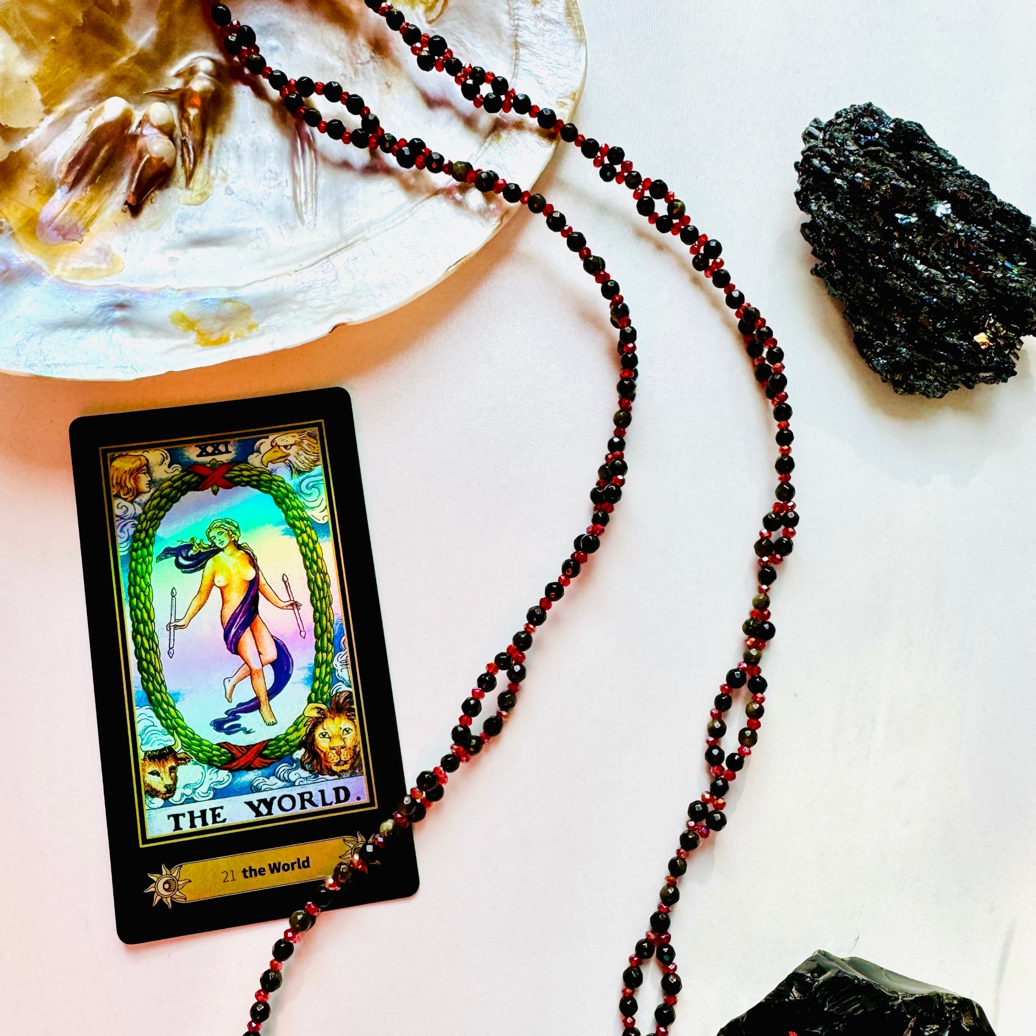 A tantric necklace with black and red beads, a yellow card with a multicolored design, and a large black obsidian stone on a white surface, with a small crystal and a feather for scale.