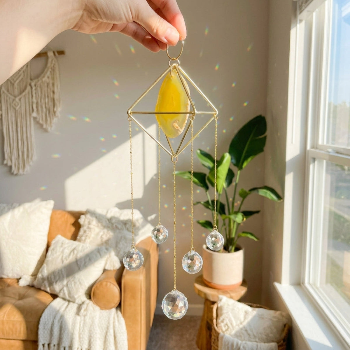A gold geometric wind spinner ornament with a yellow crystal center and clear hanging glass spheres, held by a hand in a bright, sunlit room with a couch, plant, and window.