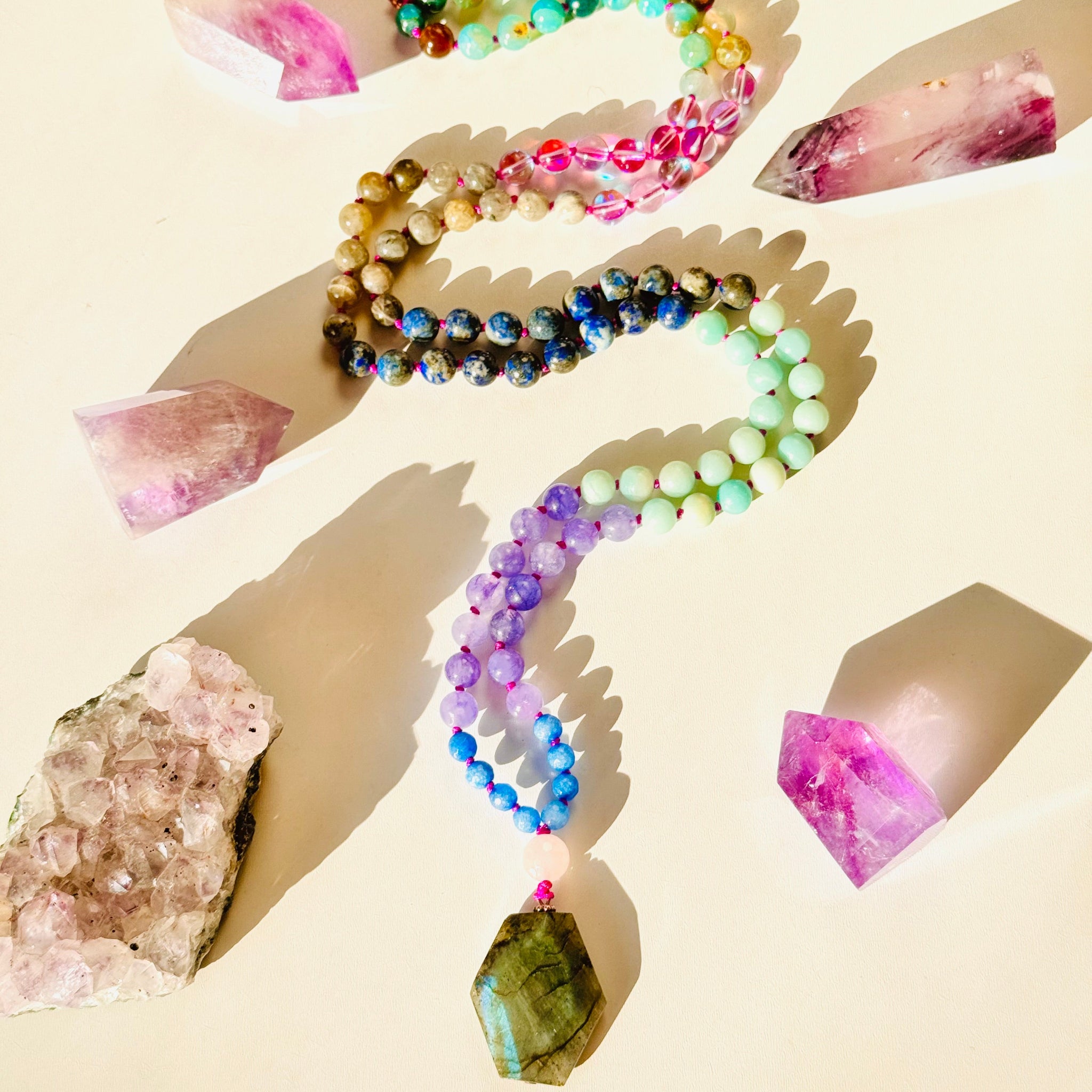 A colorful gemstone mala laid out on a light background, surrounded by raw crystals on both sides.