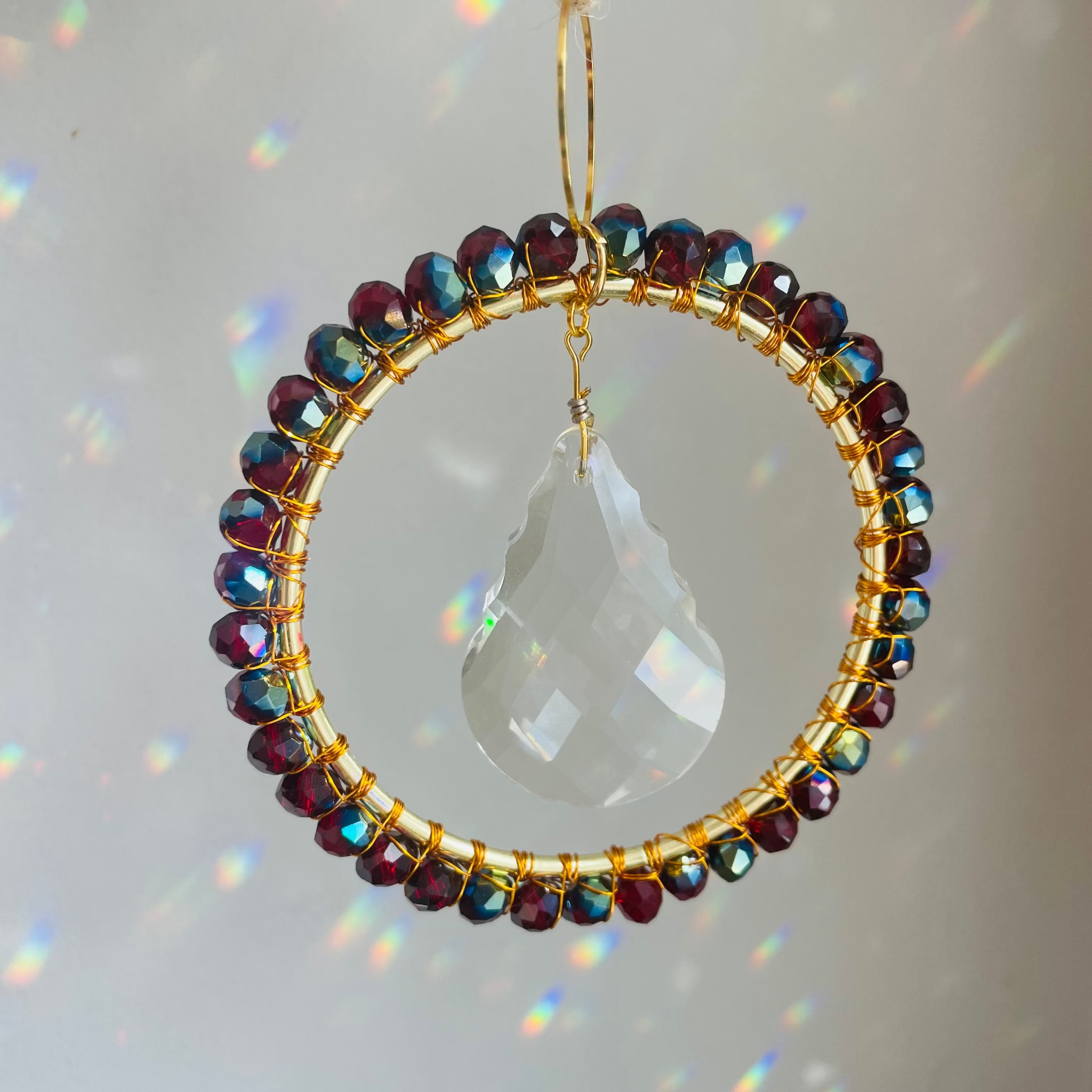 A radiant suncatcher featuring a gold-colored metal frame with blue and pink beads, and a large clear crystal in the center, hanging by a twine against a sunlit background.