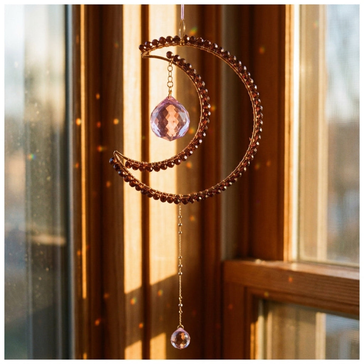 A crescent moon window suncatcher ornament hanging in front of a sunlit window. The ornament features a gold-toned crescent frame adorned with dark brown beads, a faceted pink crystal ball, and a dangling teardrop crystal charm.