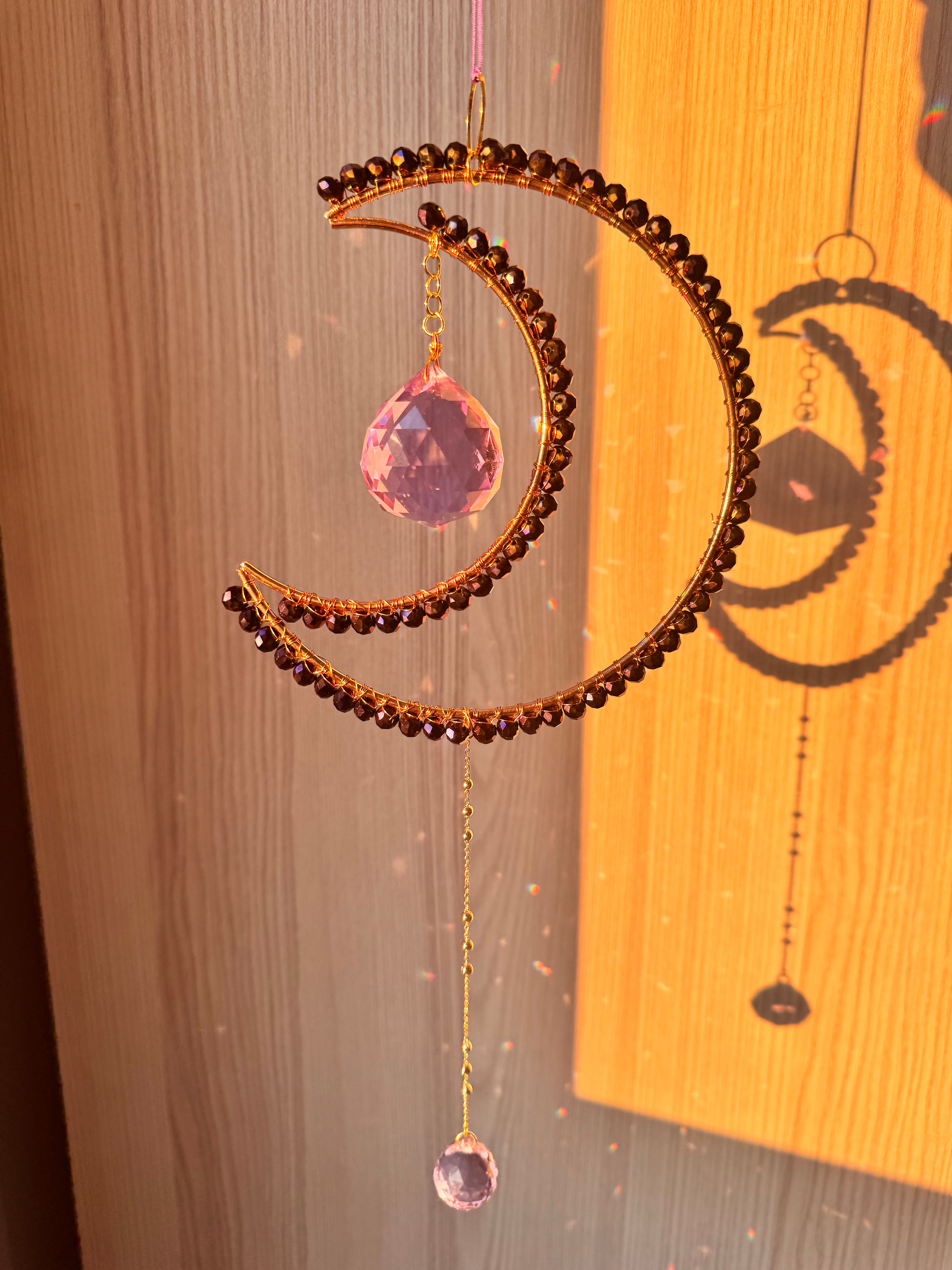 A crescent-shaped crystal suncatcher with deep indigo beads and brass frame, hanging by a string, displayed against a wooden background.