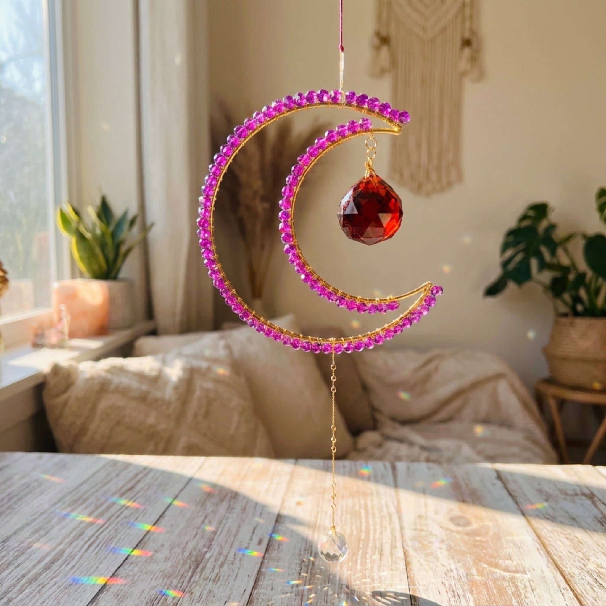 A gold crescent moon suncatcher ornament adorned with pink rhinestones and a faceted red crystal, hanging on a wooden table in a sunlit bedroom with plants and a bed in the background.