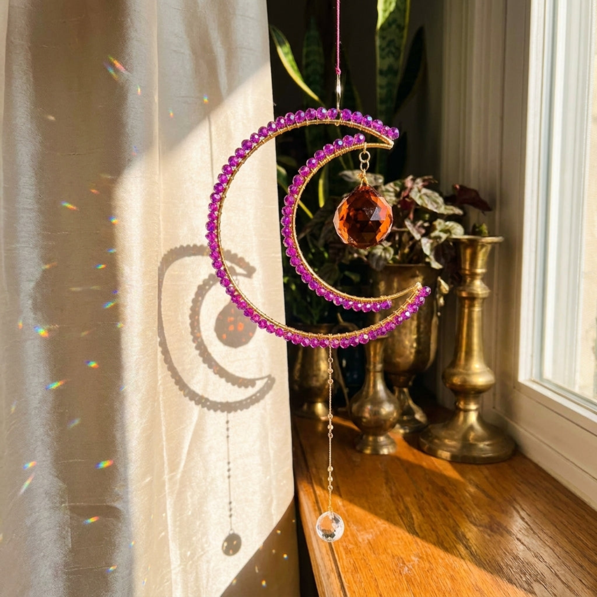 A crescent moon suncatcher with purple faceted beads and a clear hanging crystal, displayed on a window sill in a sunlit room.