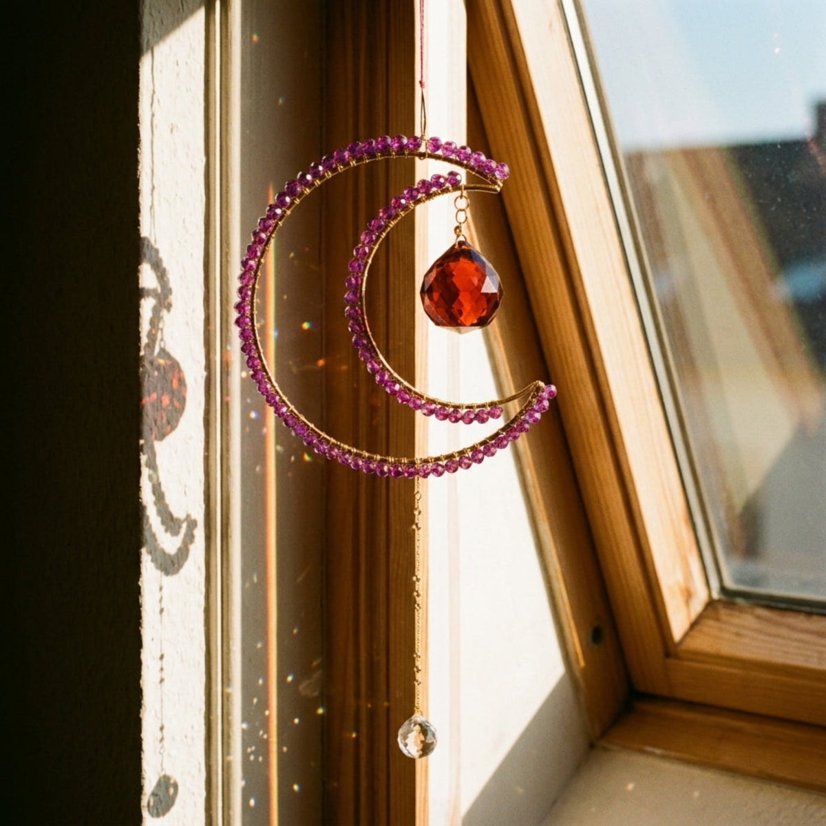 A crescent moon suncatcher ornament with pink faceted beads and a red teardrop gem, hanging in front of a wooden window with warm sunlight.