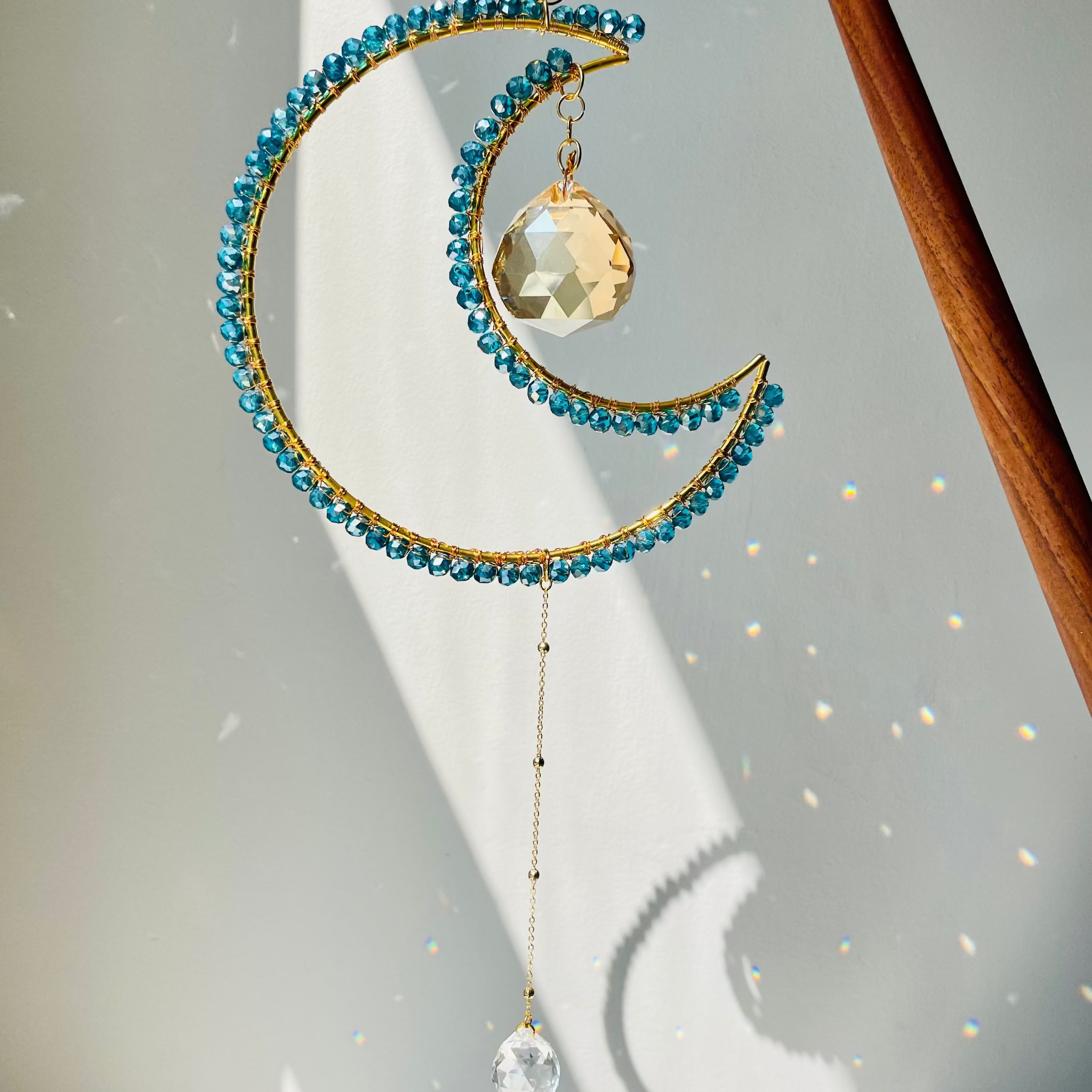 A crescent-shaped suncatcher with blue beads and a crystal pendant, hanging by a chain, with light reflecting off the beads.