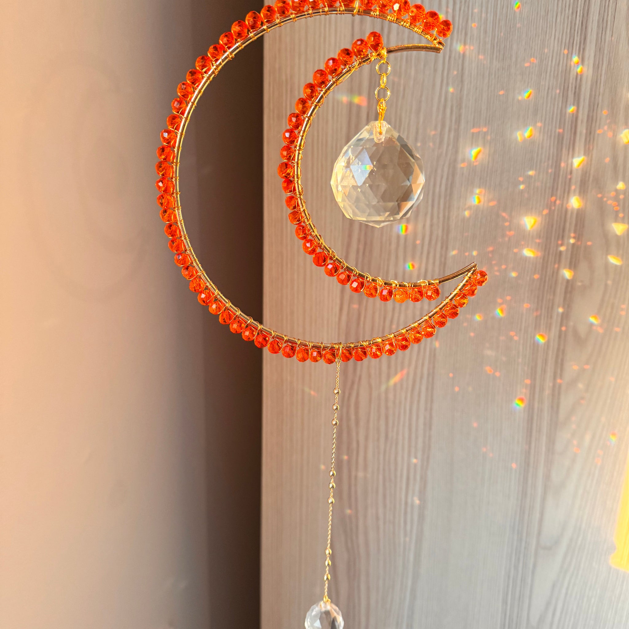 A crescent-shaped orange suncatcher with wire-wrapped beads and a crystal prism hanging against a window with light shining through, creating a colorful refraction.