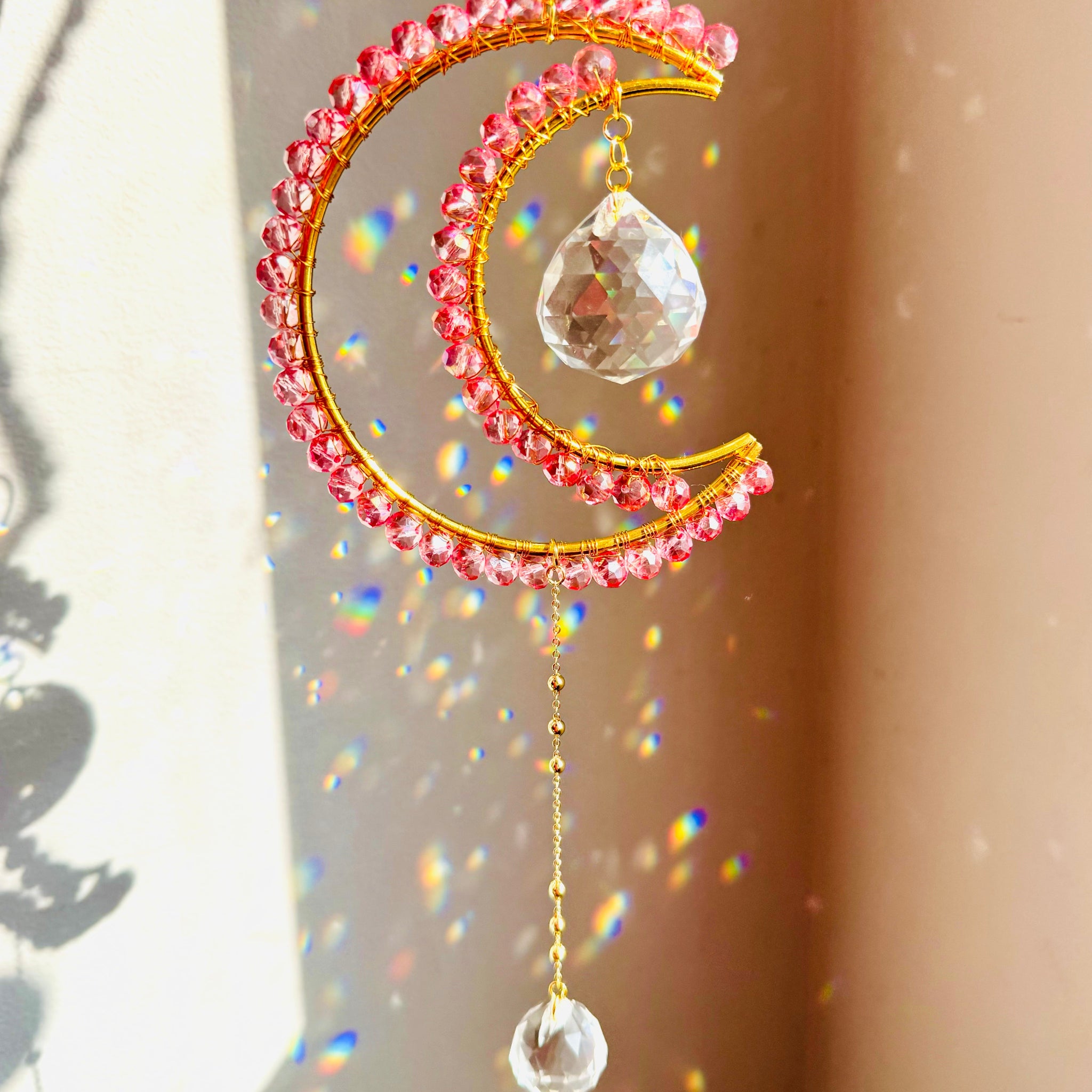 A crescent-shaped suncatcher with pink crystals and a central clear prism, hanging by a chain.