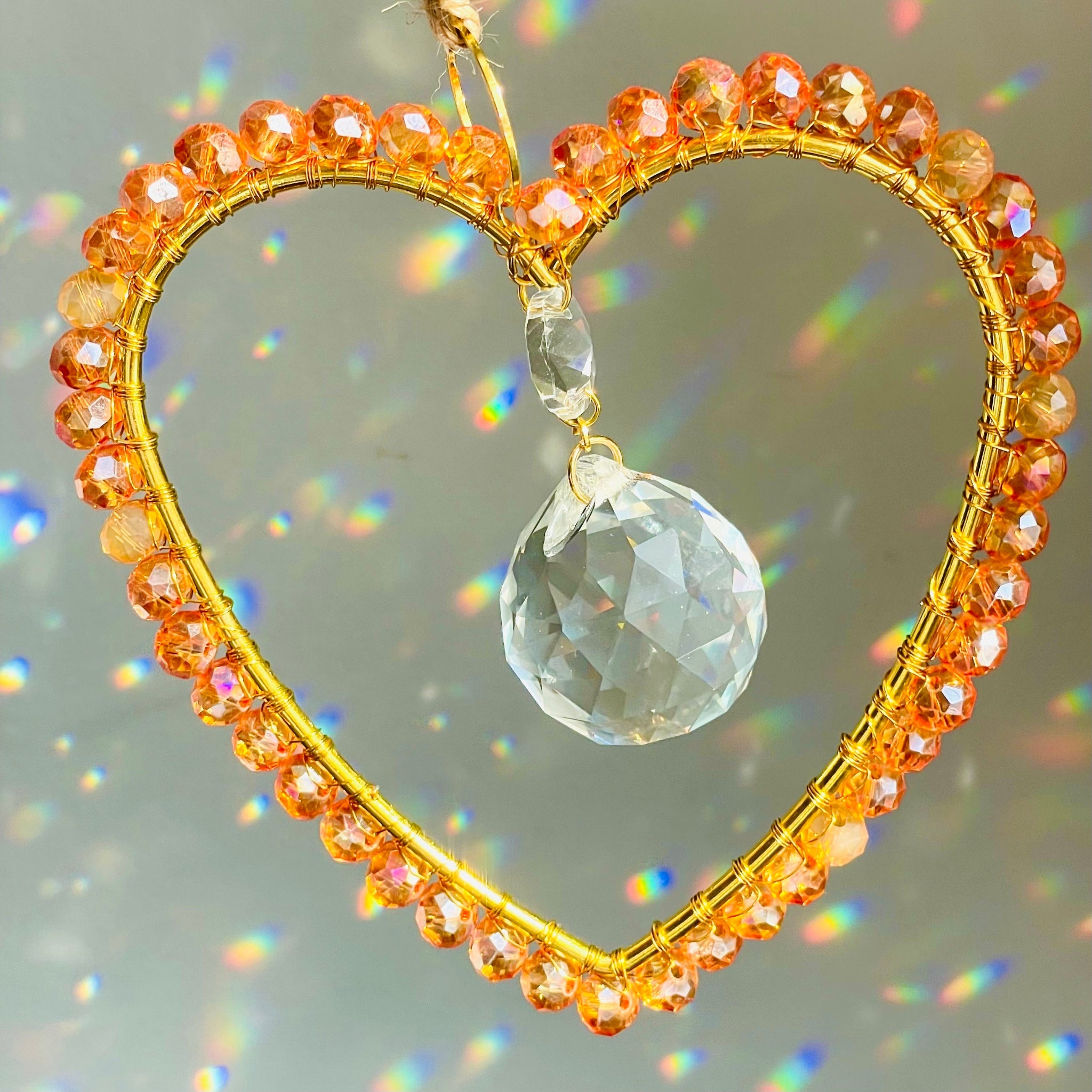 A gold-colored heart-shaped suncatcher with wire-wrapped Czech crystals arranged around a faceted glass prism, hanging by a string against a plain background.