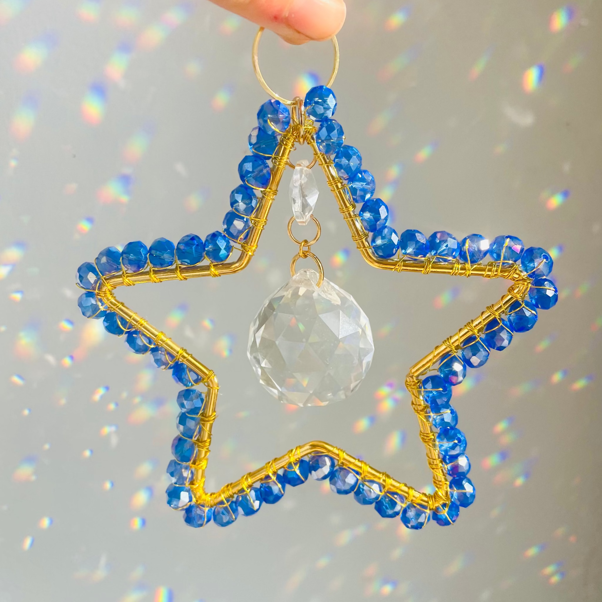 A hand holding a blue and clear crystal suncatcher in the shape of a star, with intricate gold detailing and a central pane.