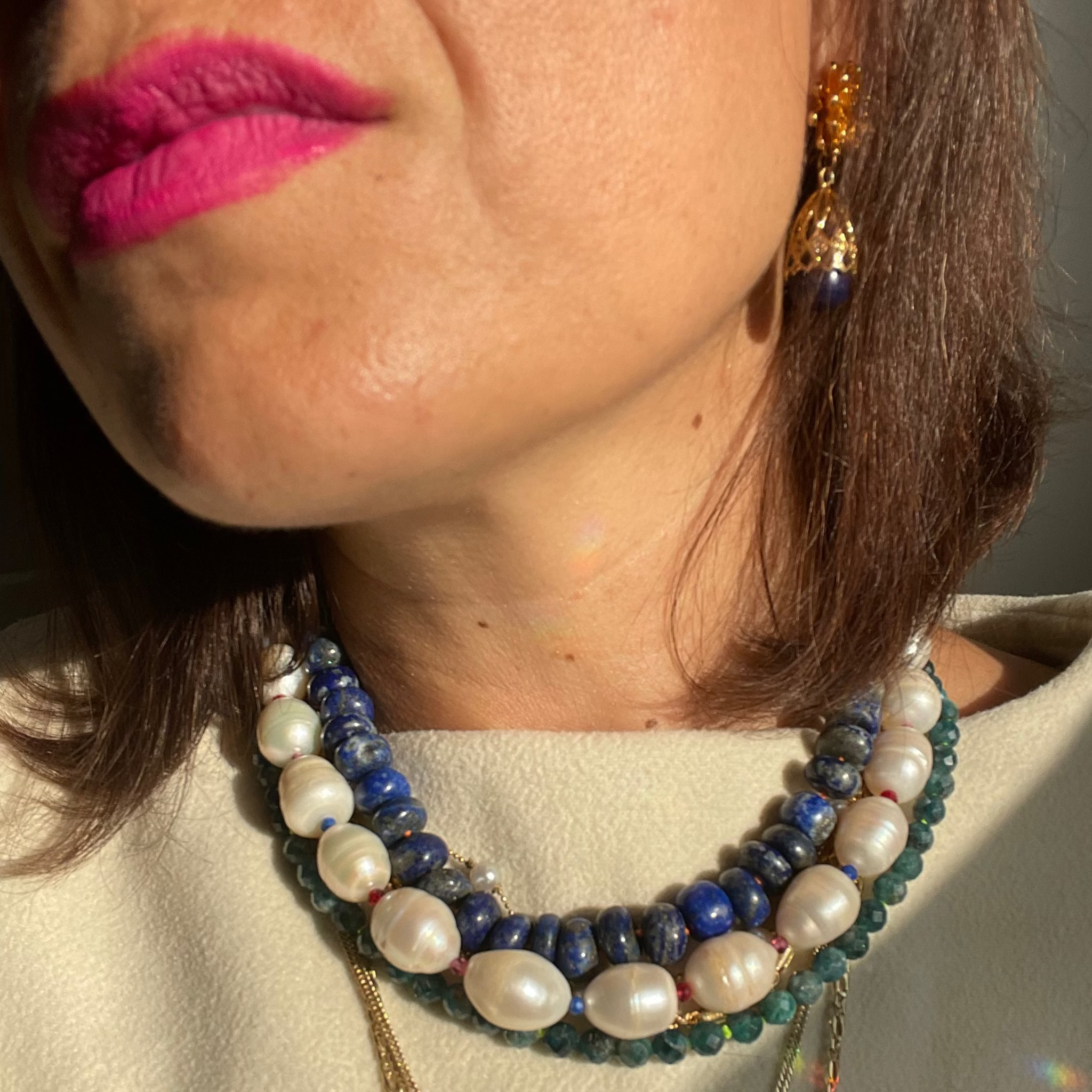 Close-up of a person wearing multiple necklaces with beads and a gold chain, with a neutral background.