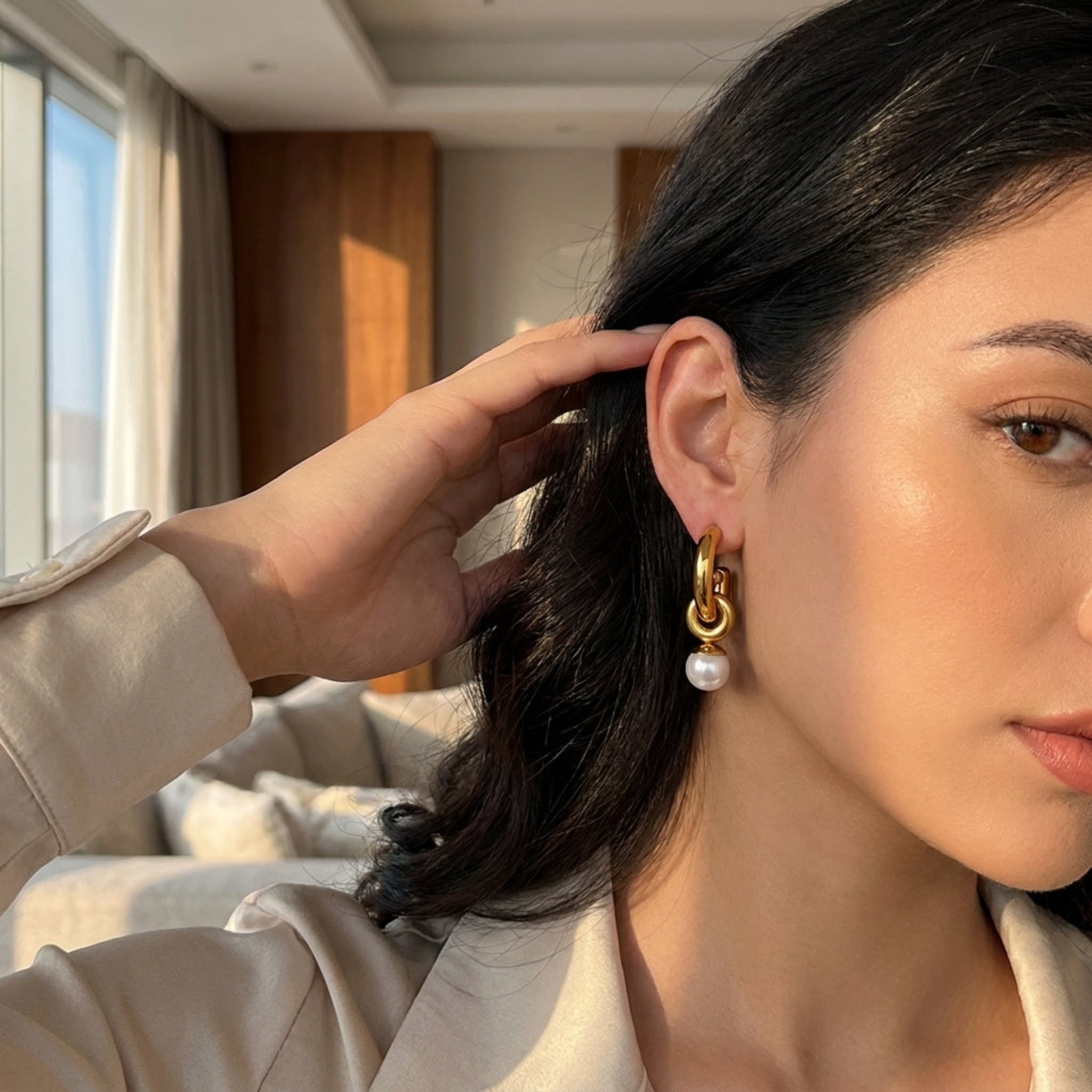 A close-up lifestyle shot of a woman wearing a gold huggie hoop earring with a dangling white pearl, set against an indoor background.