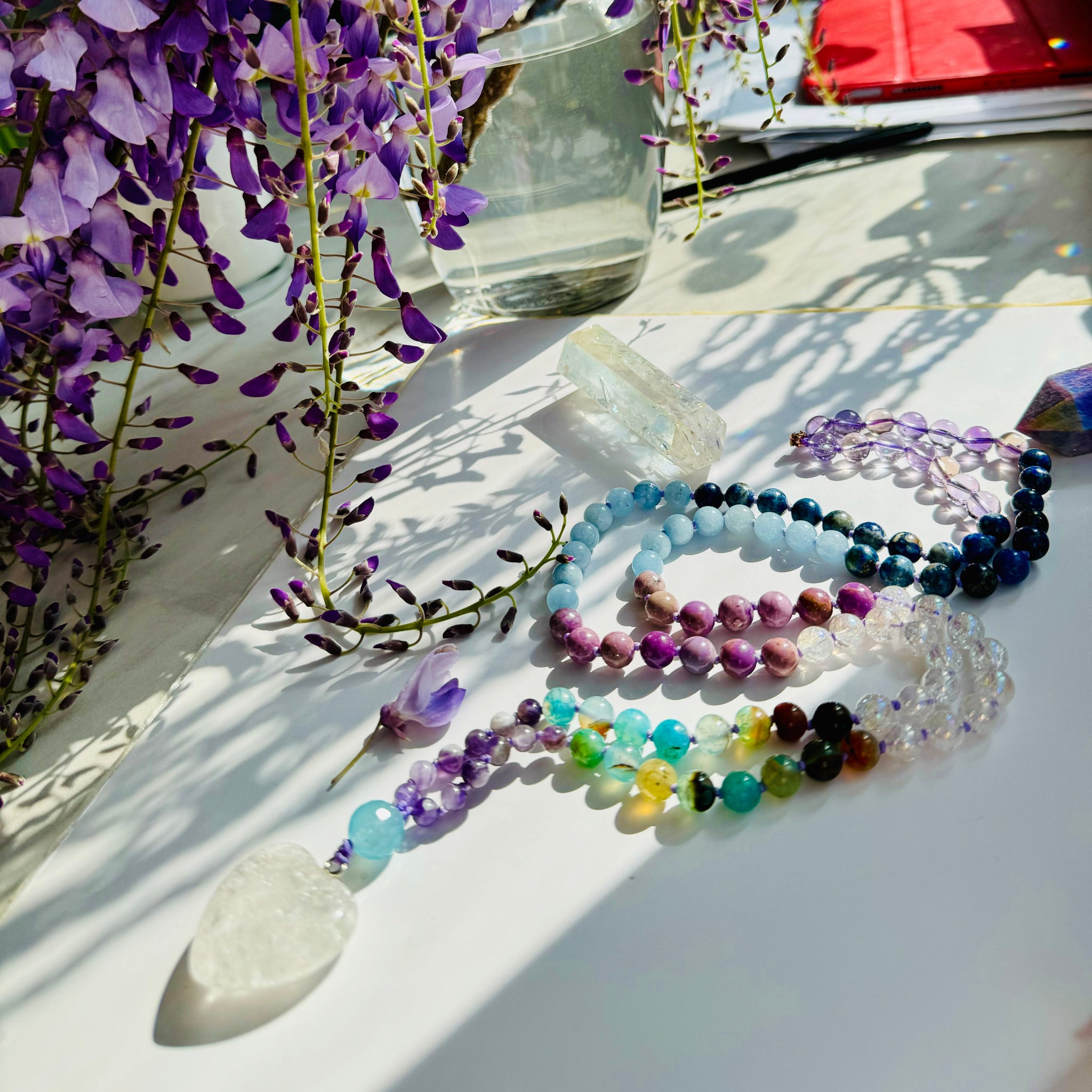 Beaded necklace with blue,purple and white crystals with white crystal heart pendant on white surface decorated with purple flowers