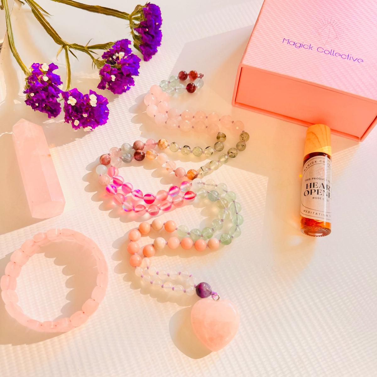 Rose Quartz mala, bracelet, tower gemstone, Magick Collective box and heart opener oil on white surface with purple flowers.