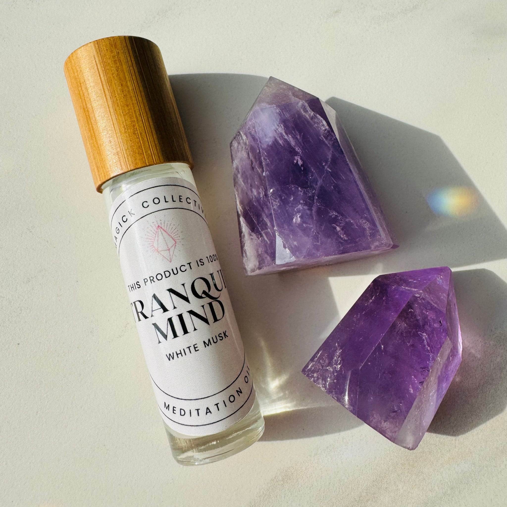A small bottle of Tranquil Mind Perfume Oil with a wooden cap, labeled with the product name and branding, alongside two pieces of polished purple amethyst gemstones.