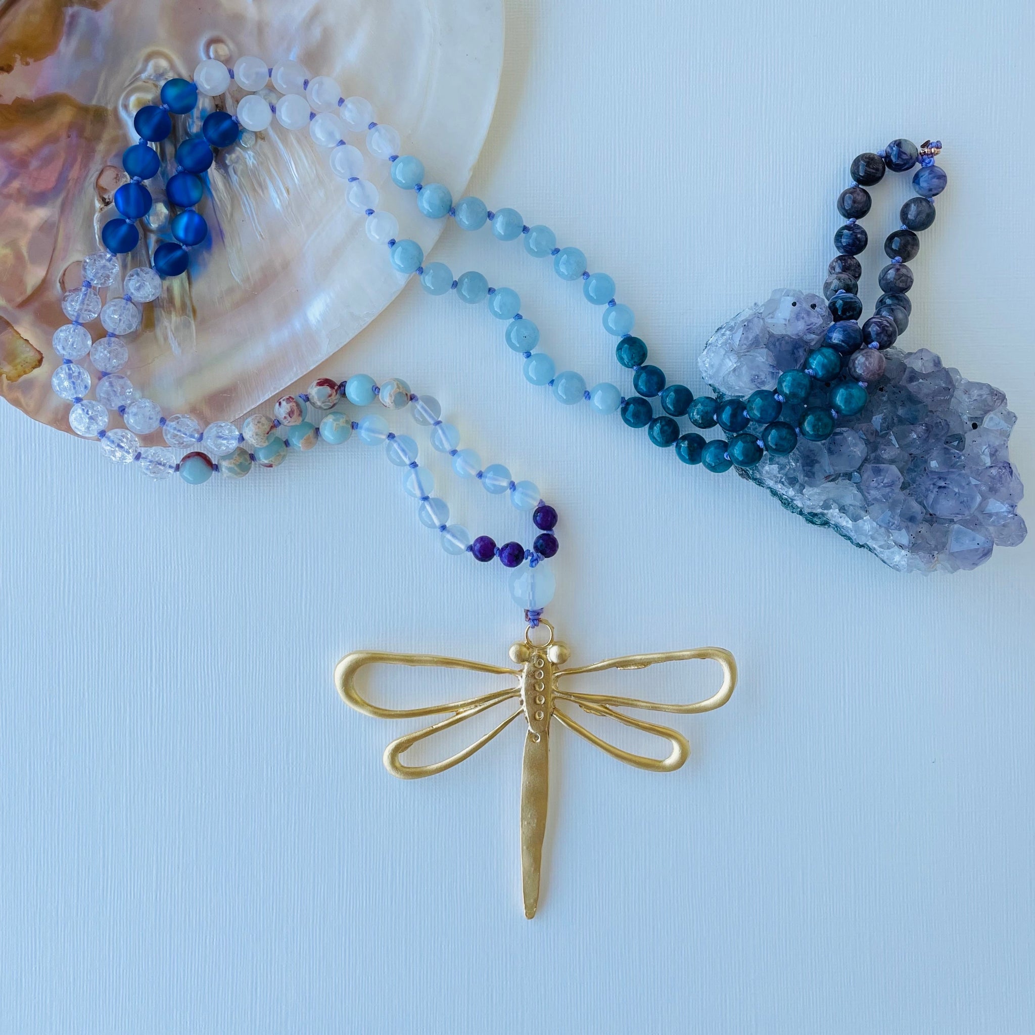 Beaded gemstone necklace with gold dragonfly pendant on white surface with shell and amethyst