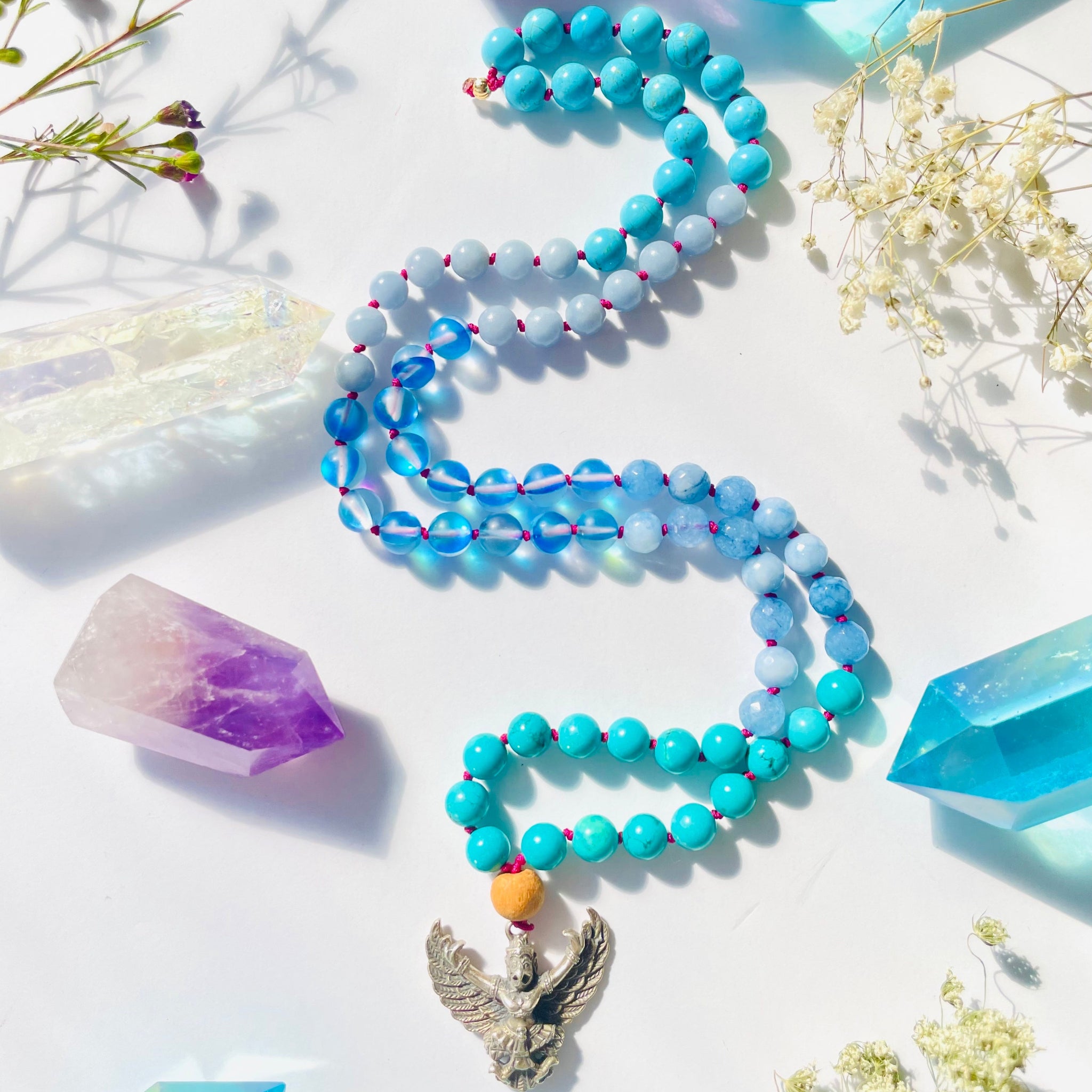 A necklace with a combination of blue gemstones and a brass garuda pendant, displayed on a white background with flowers and crystals.