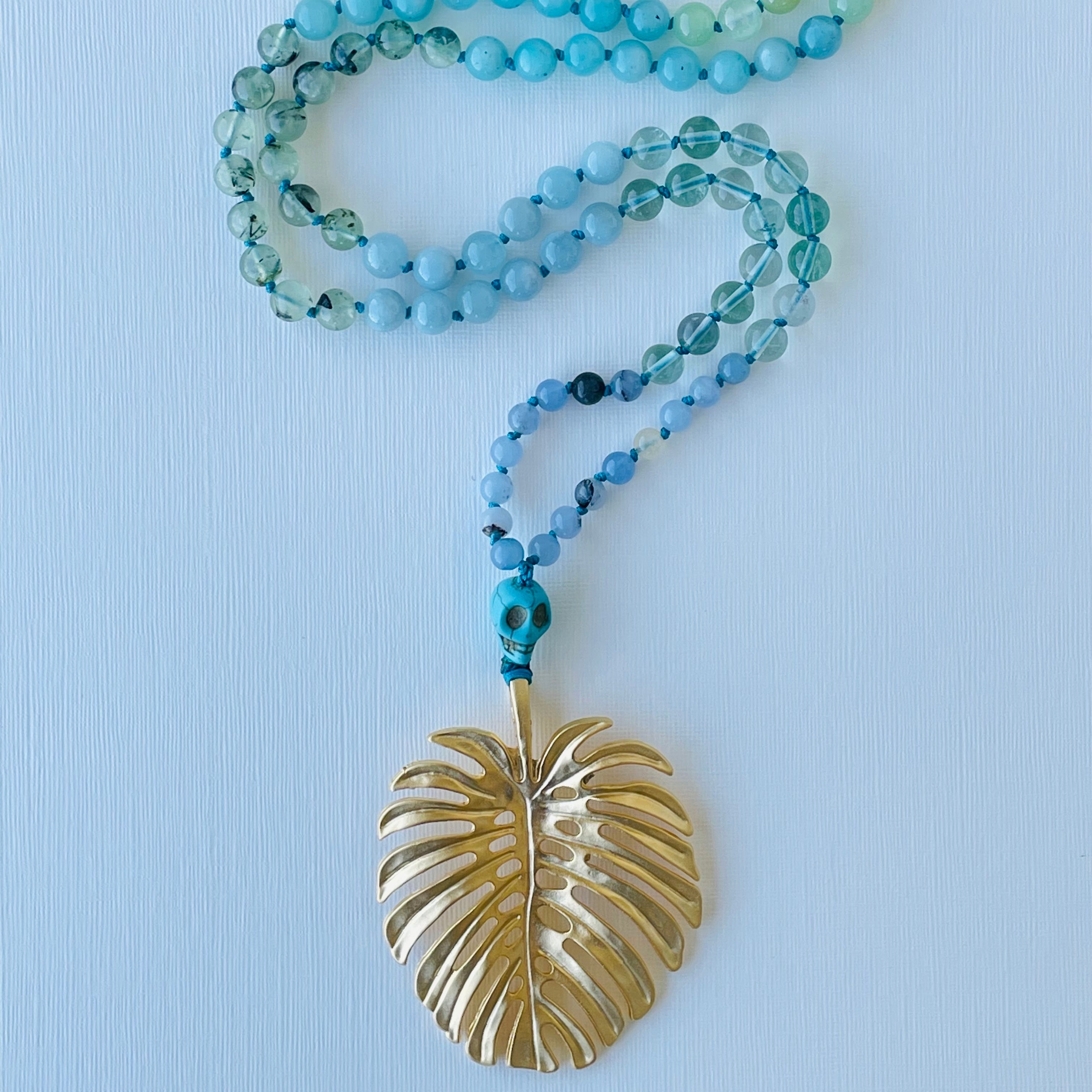 A green and blue beaded necklace with a gold-colored leaf-shaped pendant, displayed against a white background.