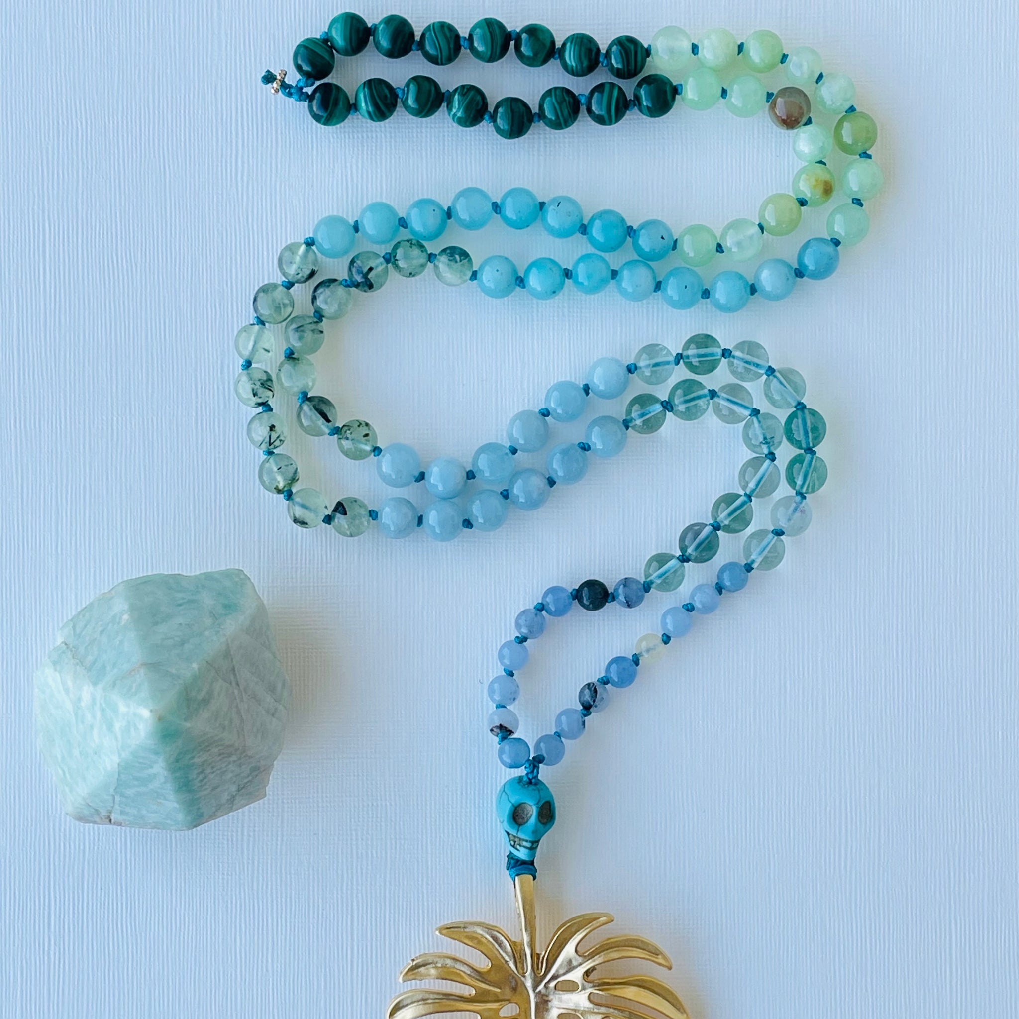 A green and blue beaded necklace with a gold-colored leaf-shaped pendant, displayed against a white background.