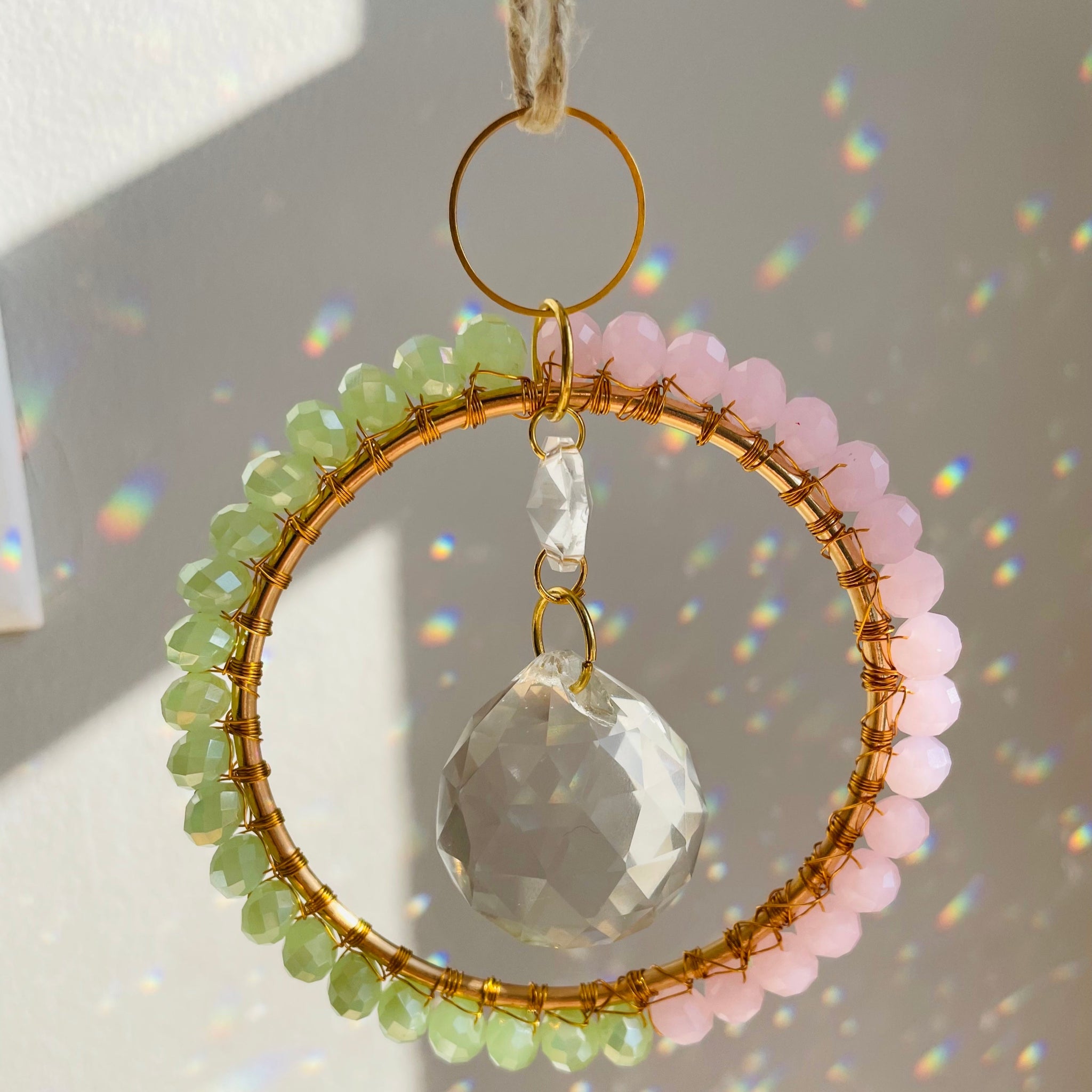 A radiant suncatcher featuring a gold-colored metal frame with blue and pink beads, and a large clear crystal in the center, hanging by a twine against a sunlit background.