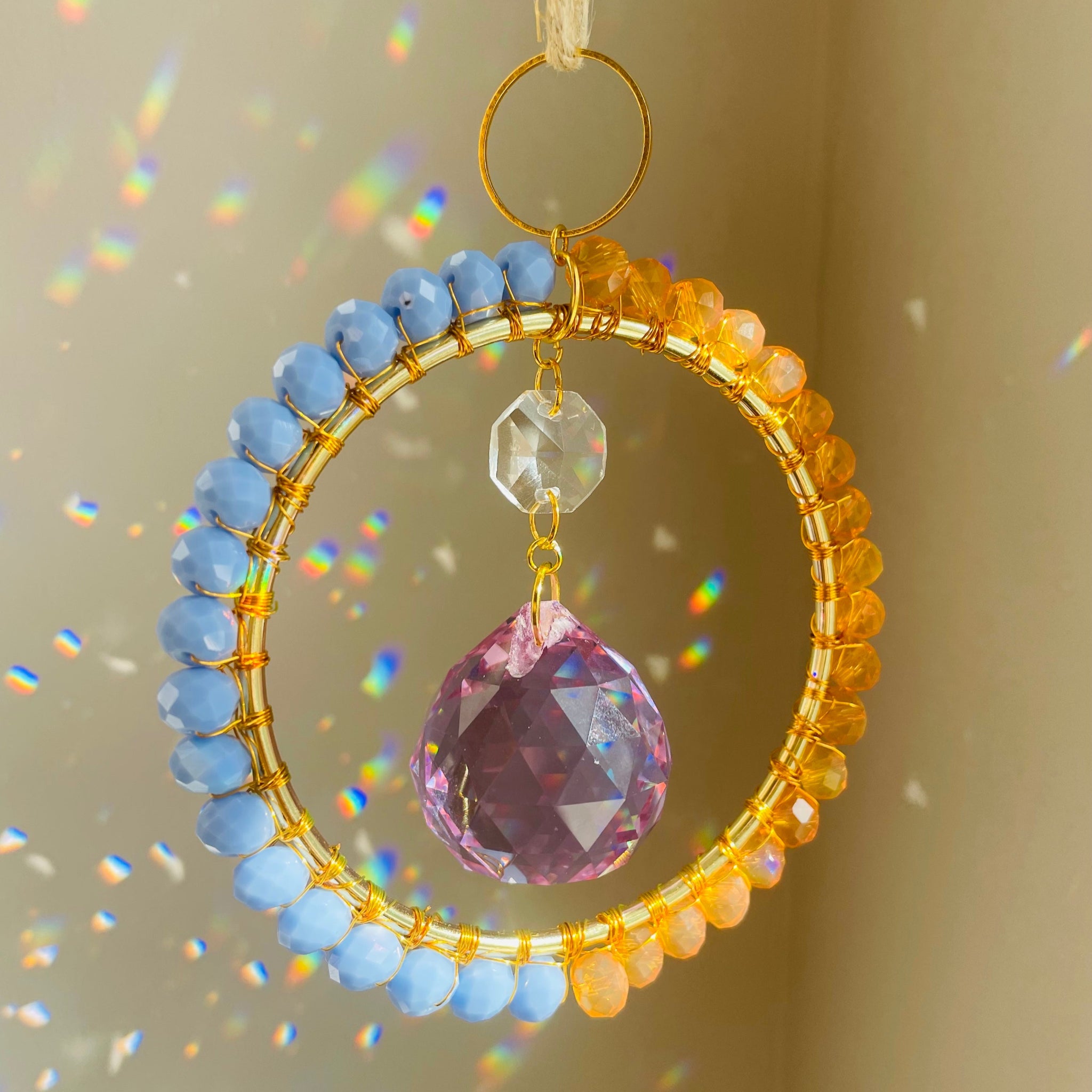 A radiant suncatcher featuring a gold-colored metal frame with blue and pink beads, and a large clear crystal in the center, hanging by a twine against a sunlit background.