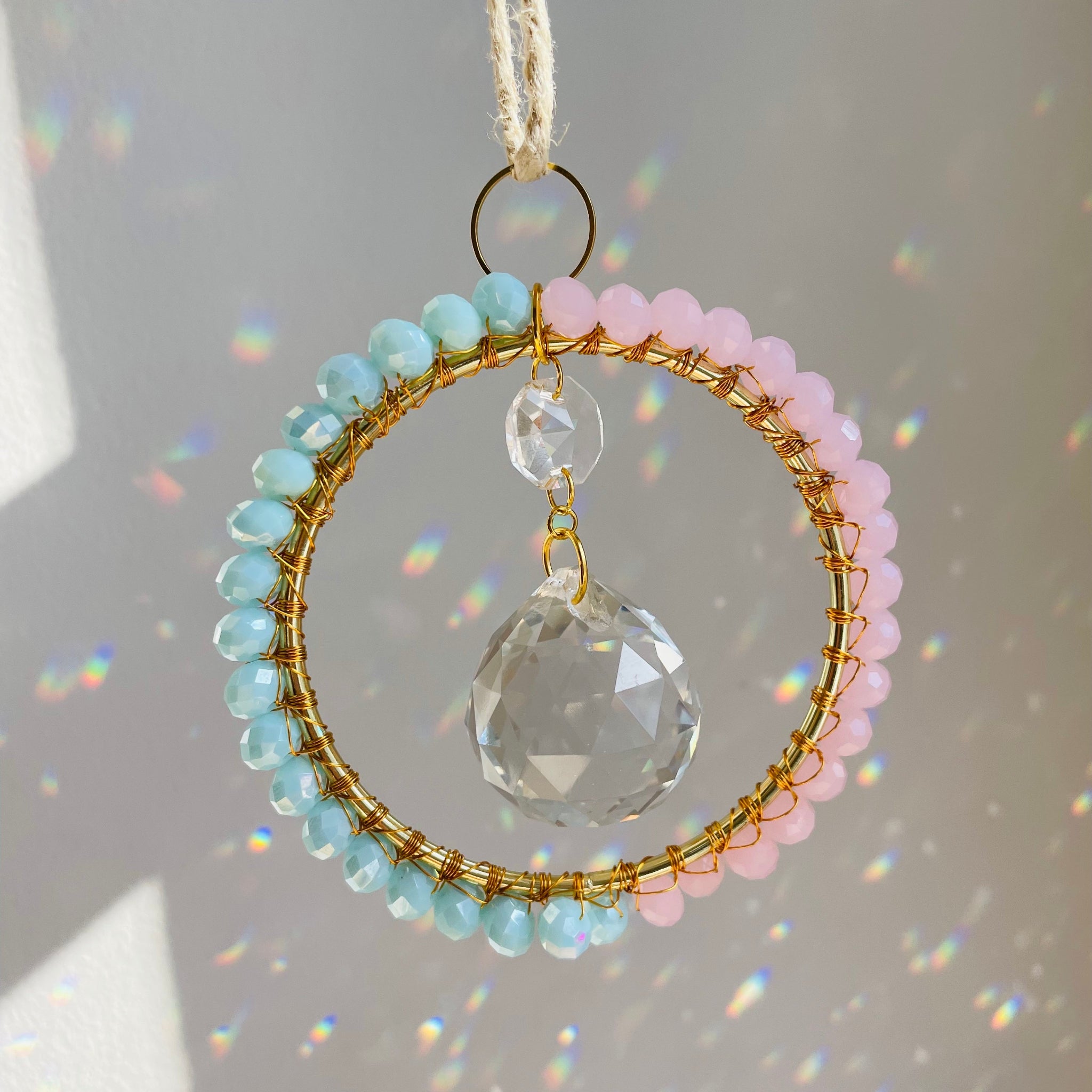 A radiant suncatcher featuring a gold-colored metal frame with blue and pink beads, and a large clear crystal in the center, hanging by a twine against a sunlit background.