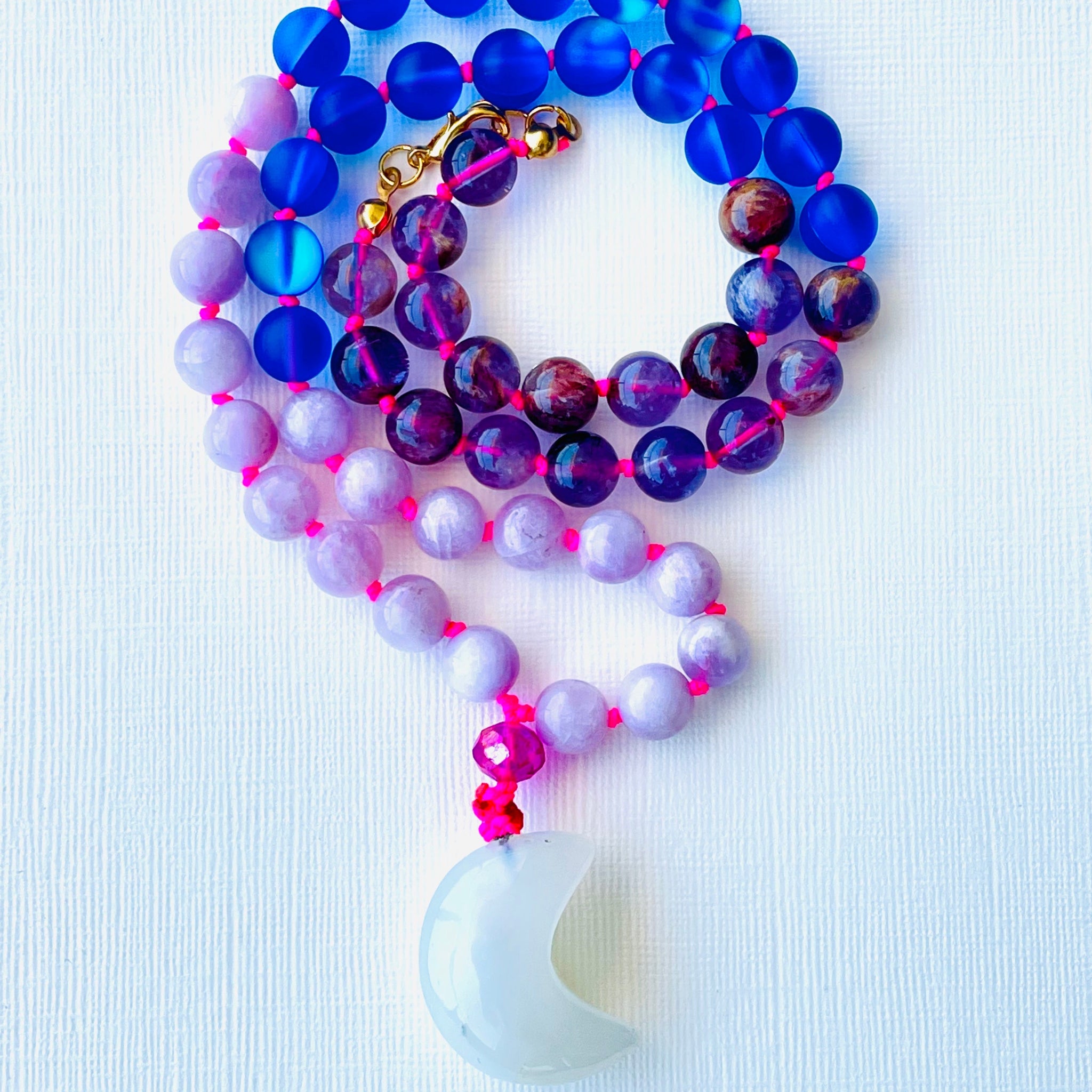 A close-up of a blue and purple gemstone mala necklace with a crescent opalite pendant, displayed on a white background.