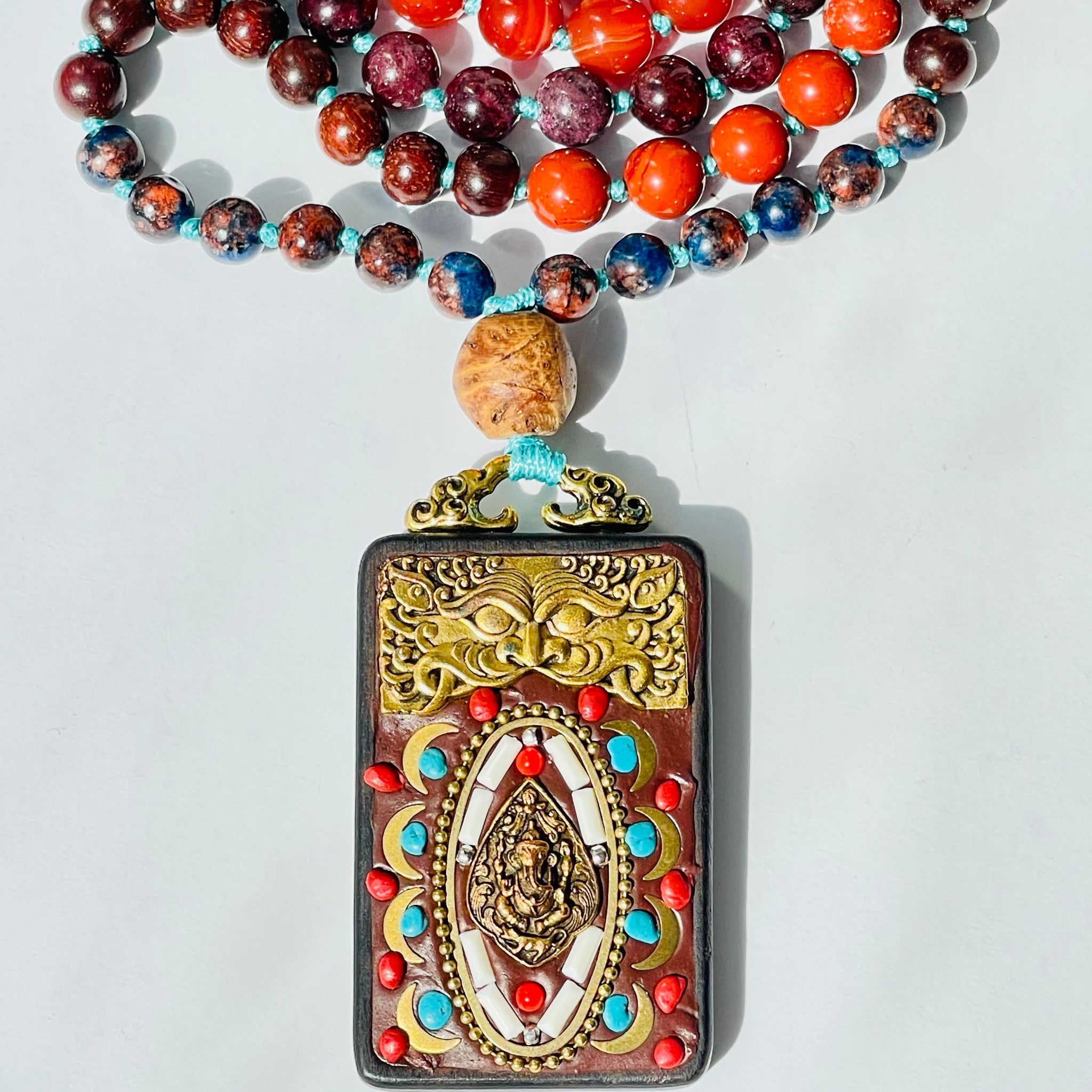 A colorful mala necklace with a mix of beads laid out on a surface with a crystal, a pendant with a deity figure, and petals scattered around.