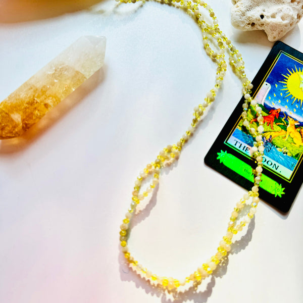 A tantric necklace with citrine gemstones, featuring a card with a sun and moon symbol, placed on a surface with a crystal, incense, and a shell.