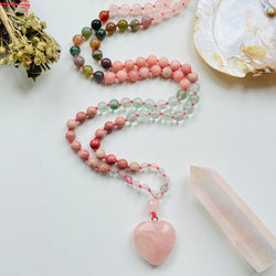 A colorful beaded mala necklace with a heart-shaped Rose Quartz pendant, displayed alongside a shell and crystals.
