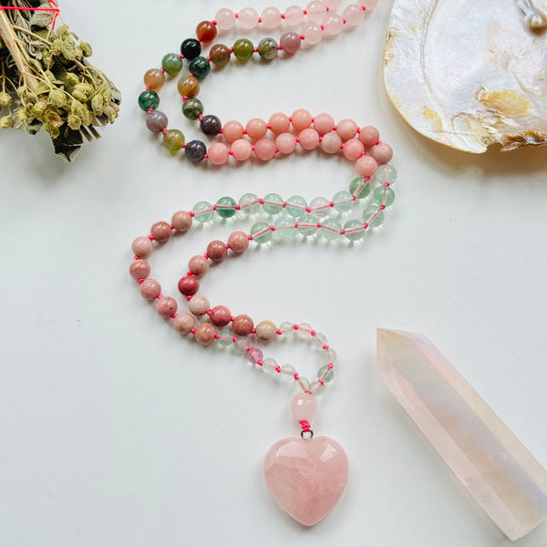 A colorful beaded mala necklace with a heart-shaped Rose Quartz pendant, displayed alongside a shell and crystals.