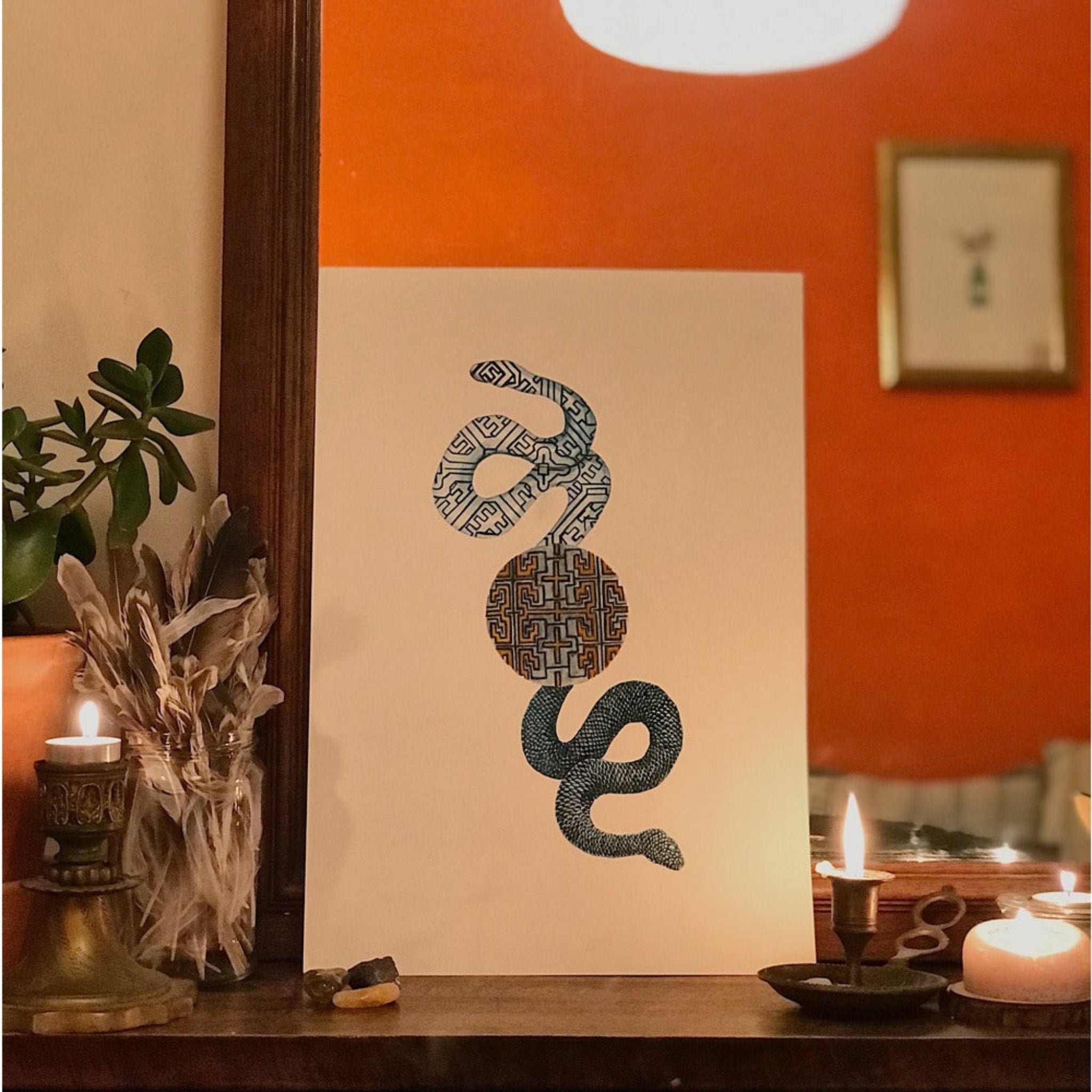 Decorative wall art with a snake design on a beige background, placed on a wooden surface with candles and plants.