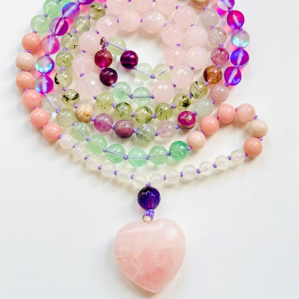 A Courageous Heart Mala necklace with a heart-shaped Rose Quartz pendant, made of various colorful beads including green, pink, purple, and white, displayed against a white background.