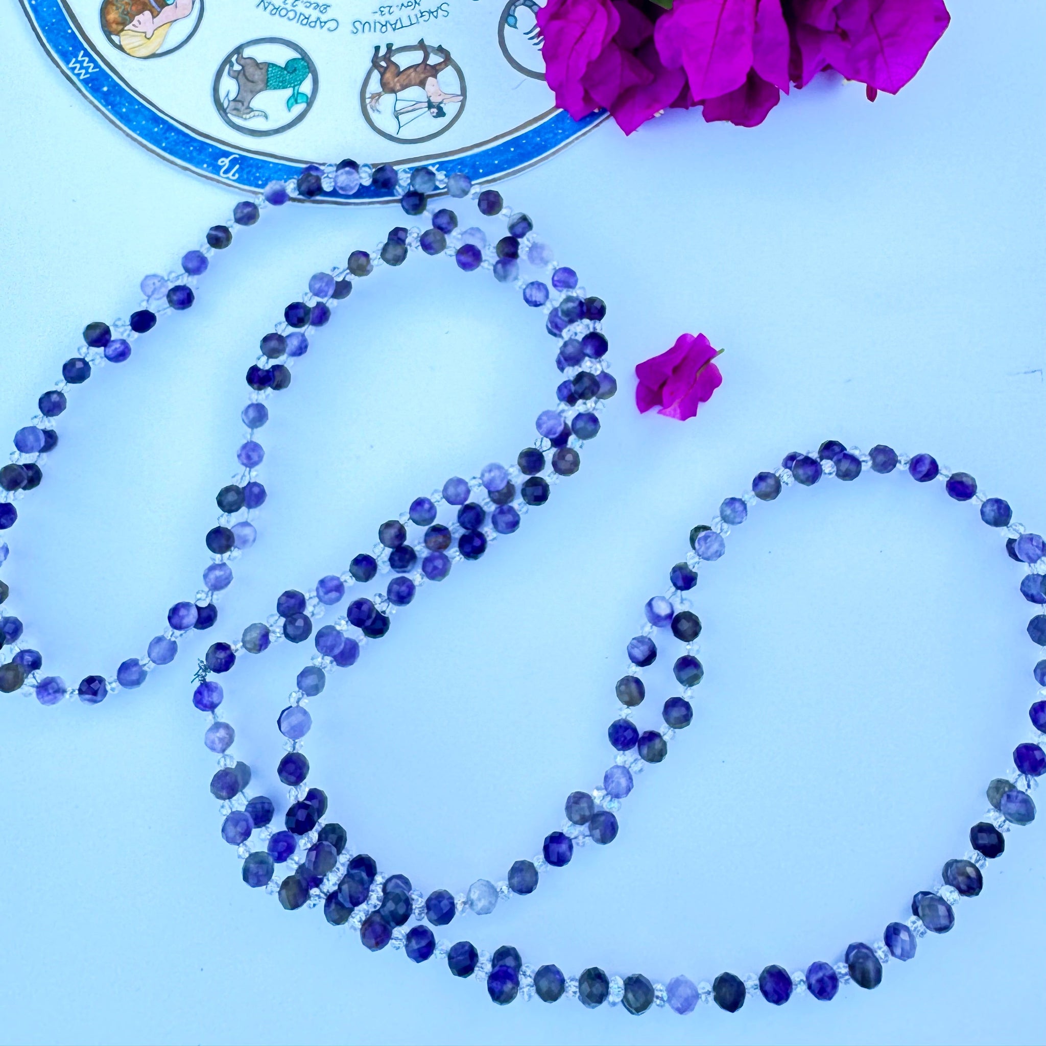 purple beaded tantric necklace on white surface with pink flowers and horoscope decor