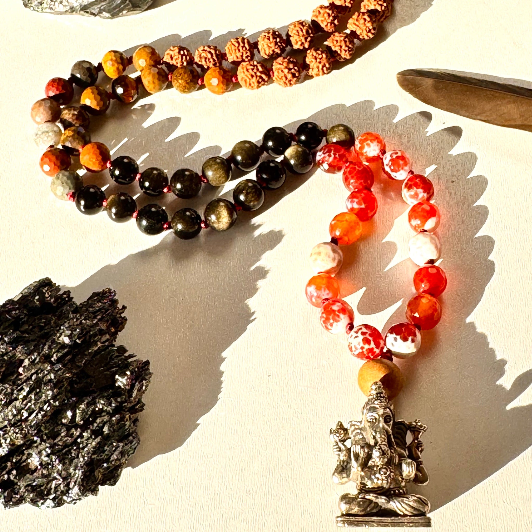 A necklace with a variety of beads including obsidian, red agate, and rudraksha, alongside a brass pendant of Ganesha. The necklace is laid out on a surface with a shadow cast over part of the beads.