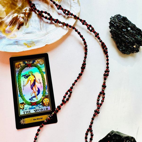 A tantric necklace with black and red beads, a yellow card with a multicolored design, and a large black obsidian stone on a white surface, with a small crystal and a feather for scale.