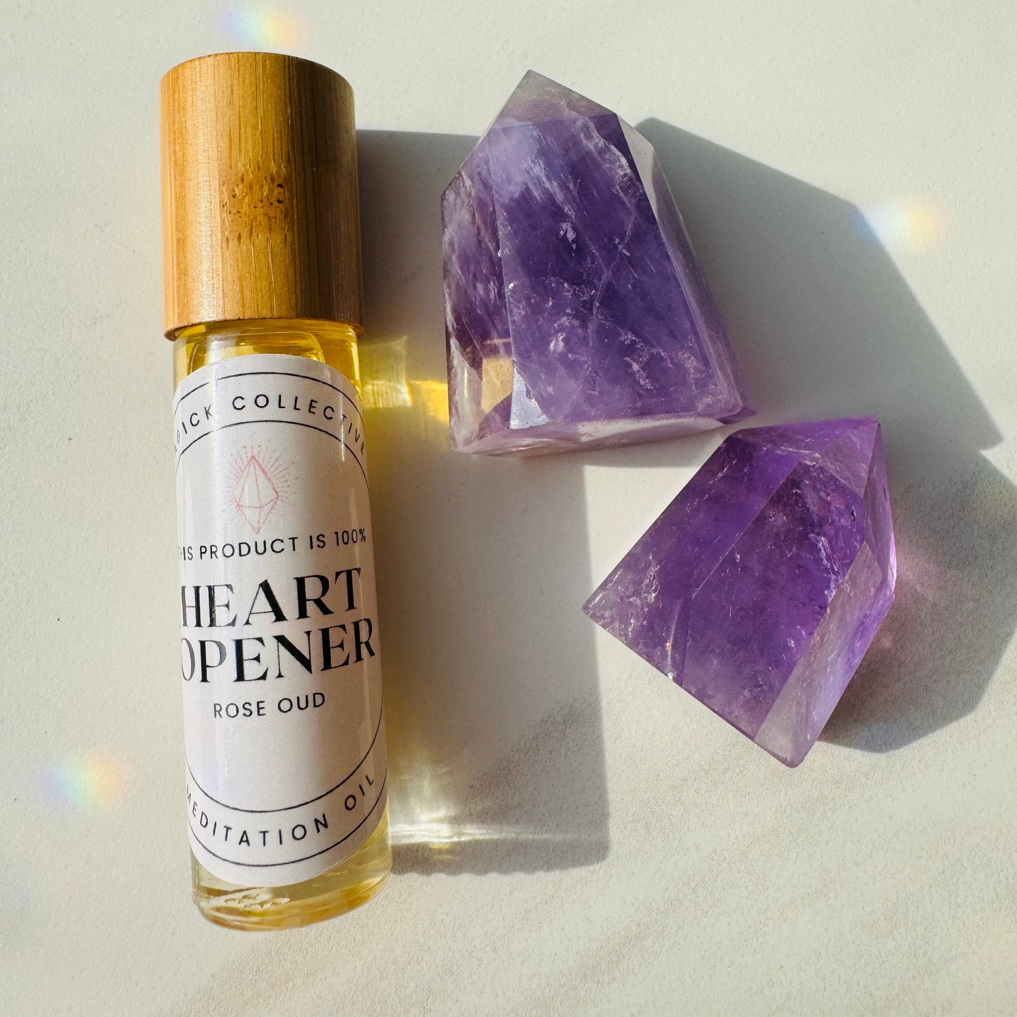 A bottle of 'Heart Opener' oil with a wooden cap, alongside a large purple crystal. Both the bottle and the crystal are placed on a reflective surface with light reflections.