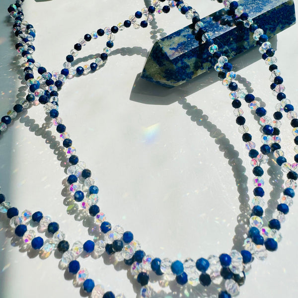 A tantric necklace with blue and clear gemstones, featuring a pendant with a blue lapis lazuli stone and a crystal wand.