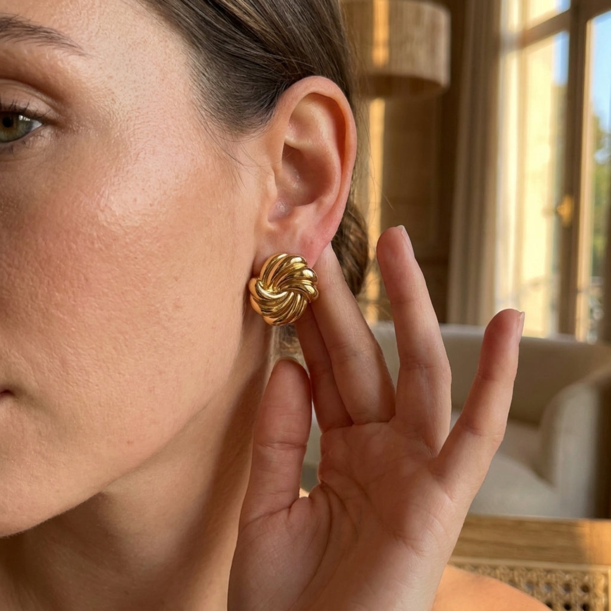 A close-up lifestyle shot of a woman wearing a textured, twisted gold earring, with her hand resting near her ear in a softly lit indoor setting.