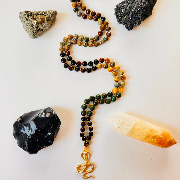 Kundalini Shakti Mala with a series of brown and multicolored beads, a brass serpent symbol pendant, and adorned with a crystal on a white background.