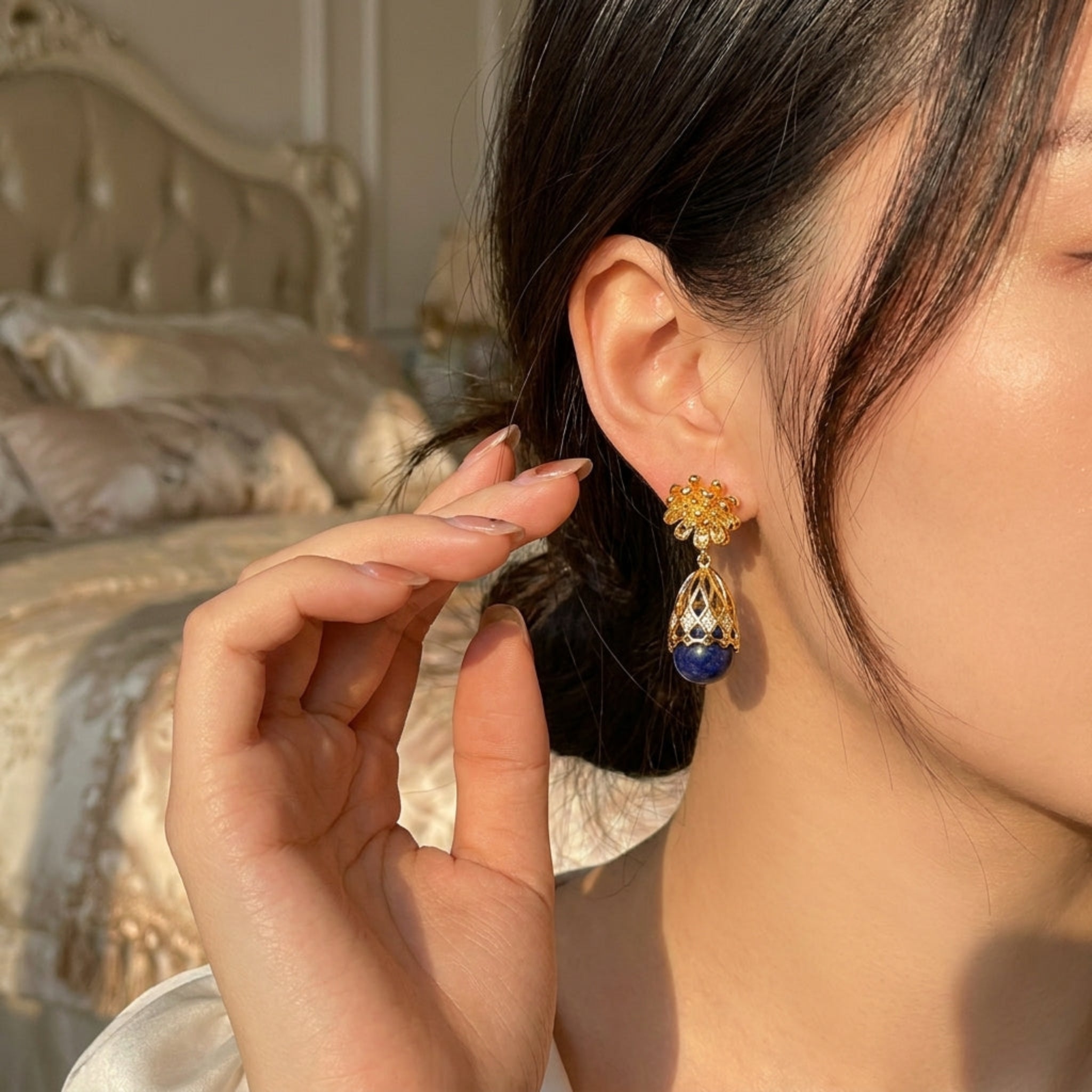 A close-up lifestyle shot of a woman wearing a gold and blue dangle earring, with her hand near her ear. The background shows a softly blurred bedroom setting with a bed and pillows.