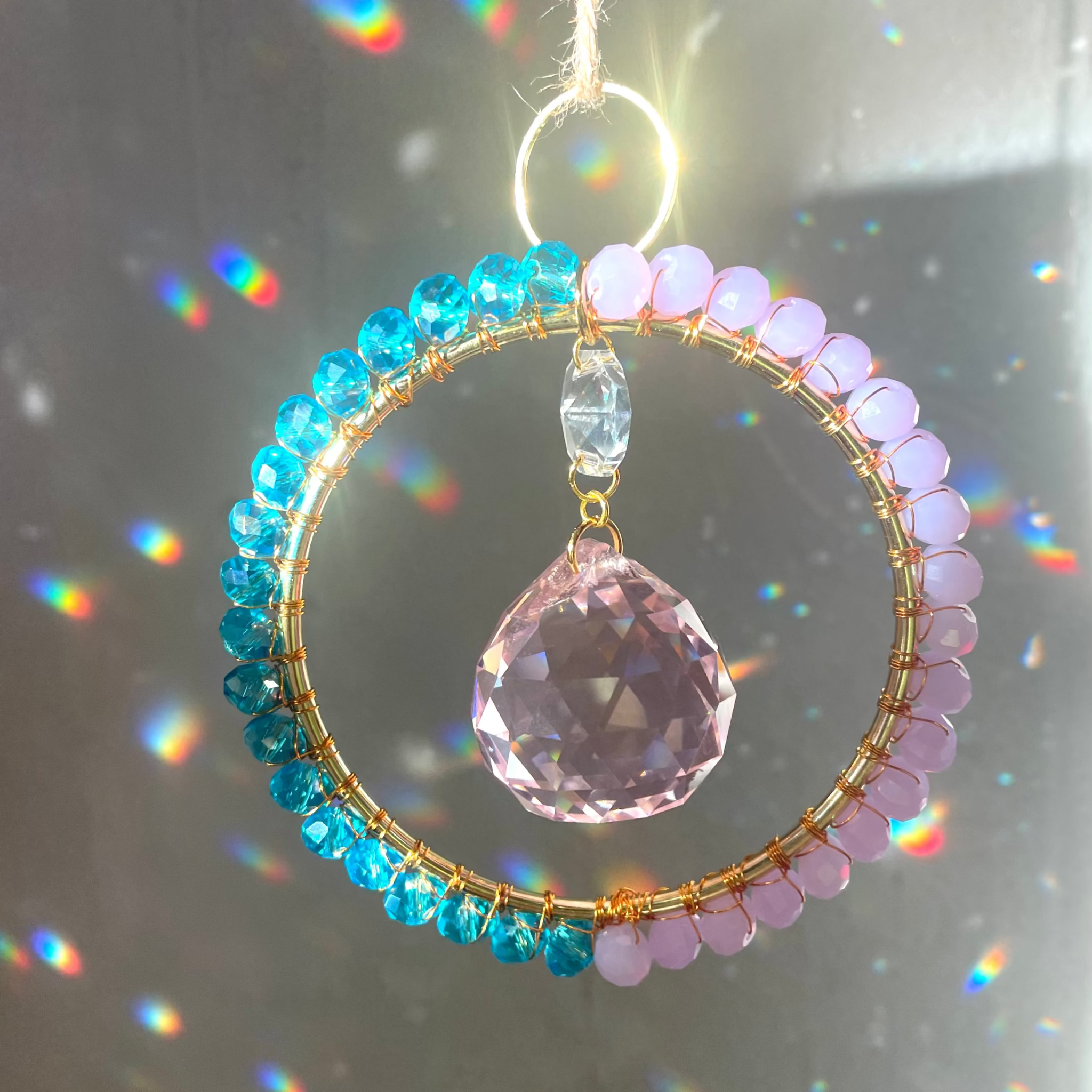 A radiant suncatcher featuring a gold-colored metal frame with blue and pink beads, and a large clear crystal in the center, hanging by a twine against a sunlit background.