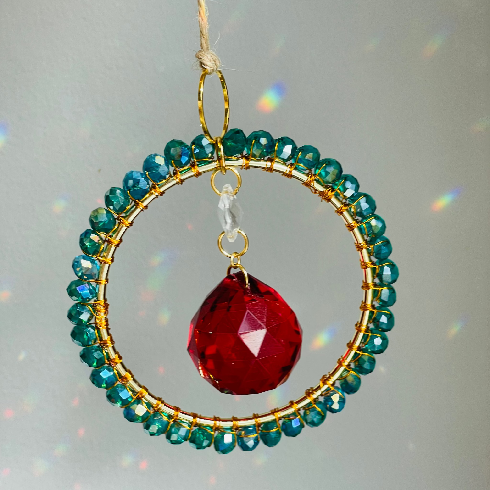 A radiant suncatcher featuring a gold-colored metal frame with blue and pink beads, and a large clear crystal in the center, hanging by a twine against a sunlit background.