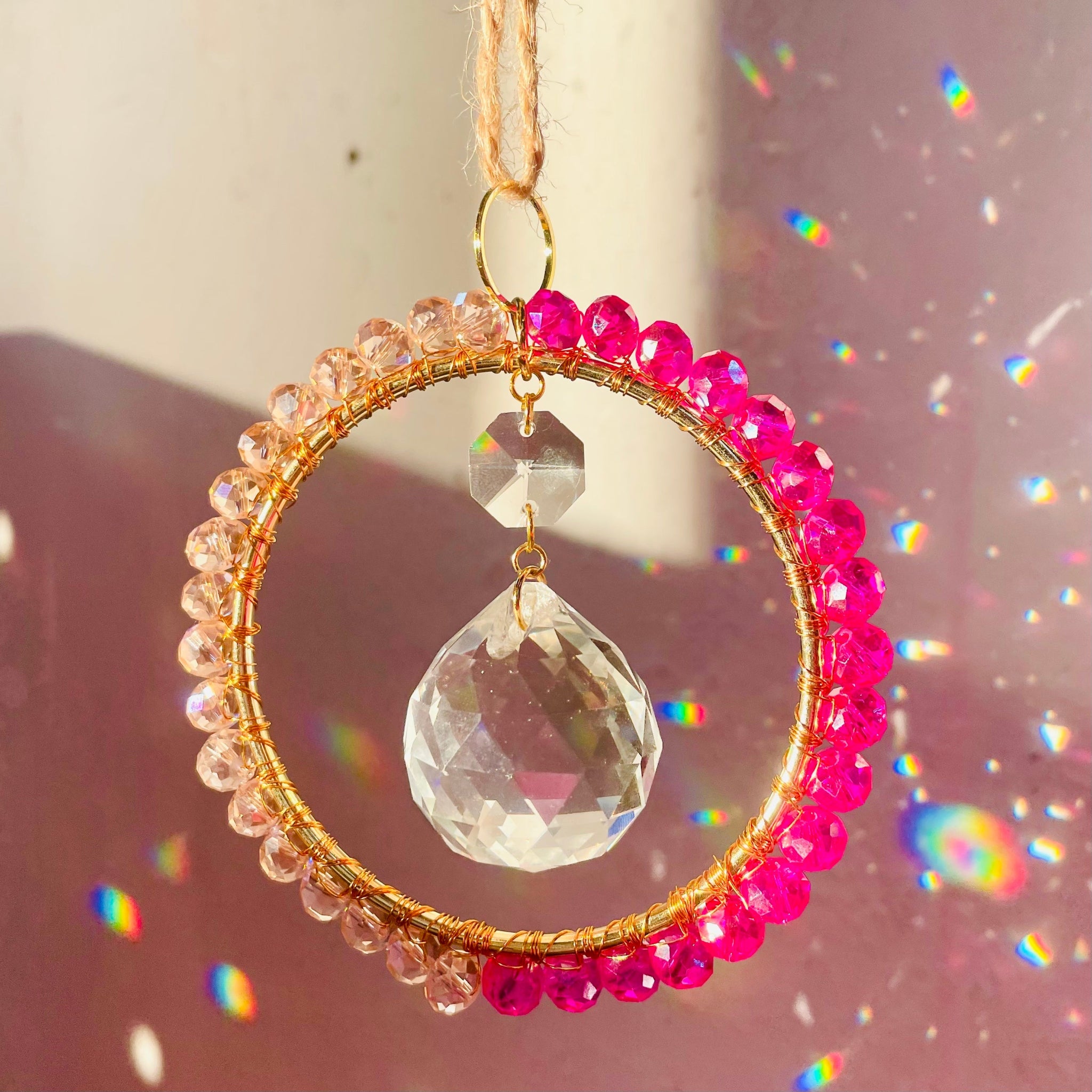 A radiant suncatcher featuring a gold-colored metal frame with blue and pink beads, and a large clear crystal in the center, hanging by a twine against a sunlit background.