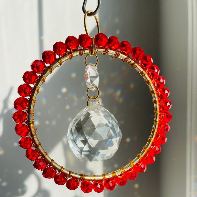A radiant suncatcher featuring a gold-colored metal frame with blue and pink beads, and a large clear crystal in the center, hanging by a twine against a sunlit background.